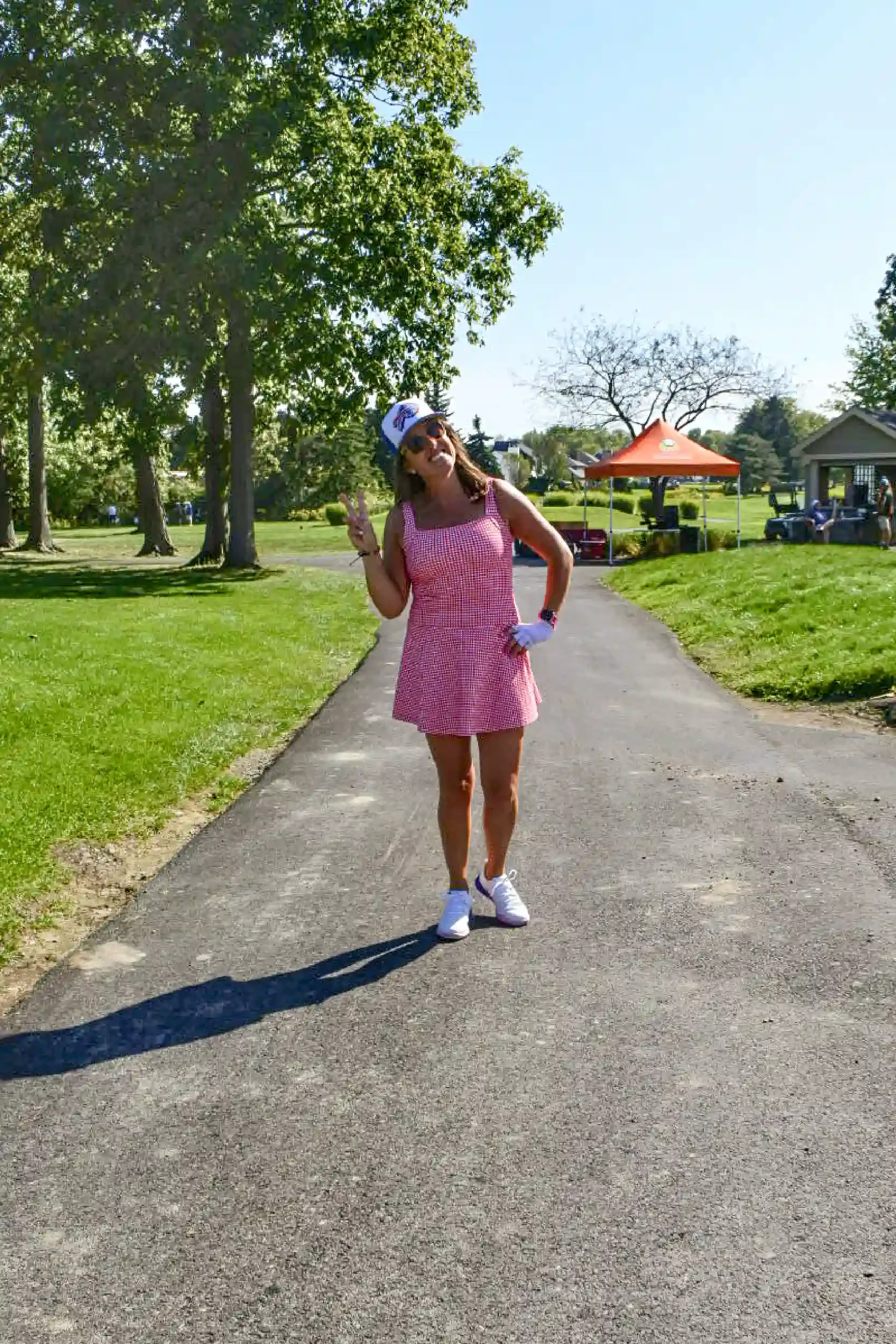 A woman wearing a pink dress, white cap, sunglasses, and white sneakers standing on a paved pathway in a park, making a peace sign with her right hand, with trees and a red canopy in the background.