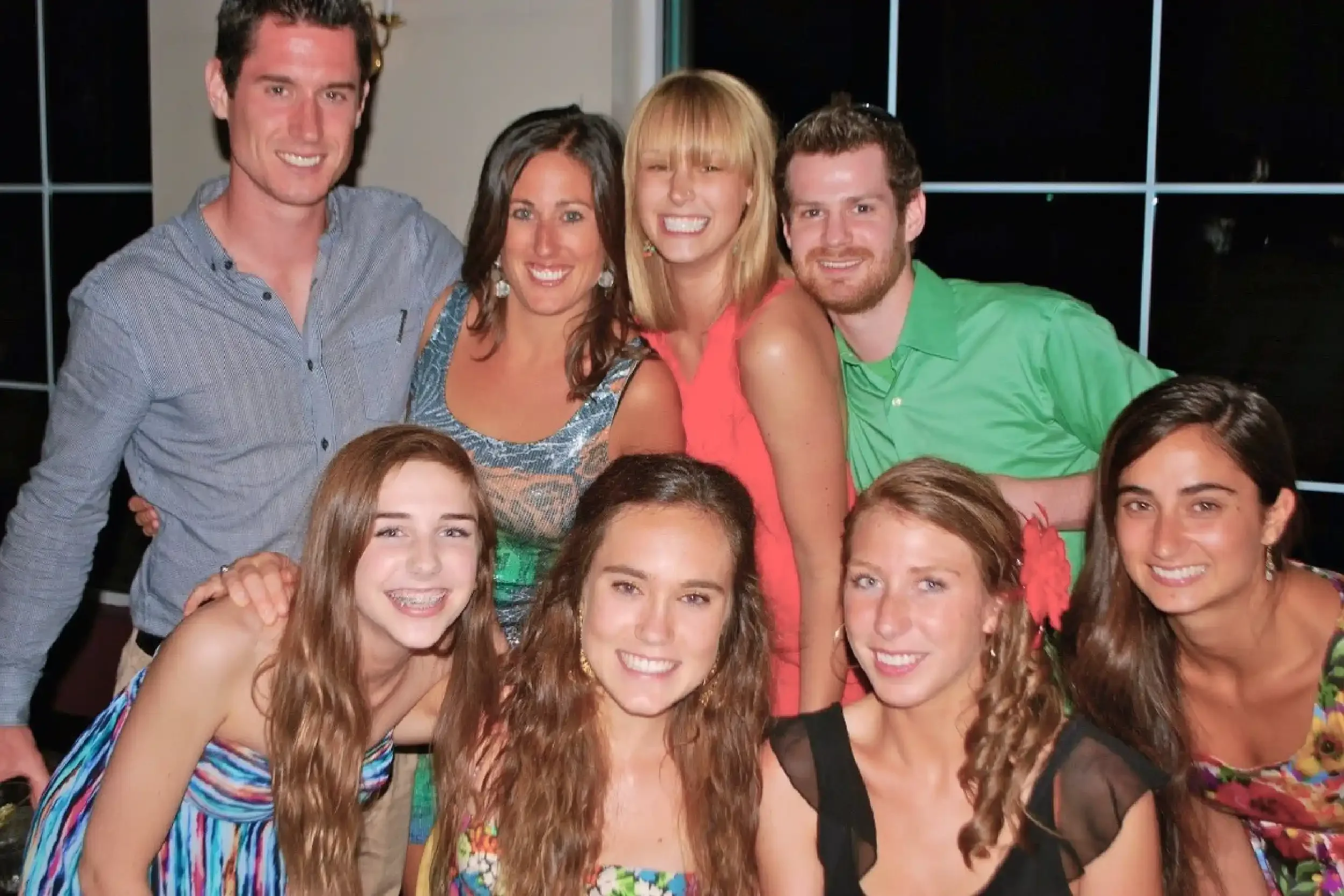 A group of smiling people, young women and men, at a social gathering, with a dark background and a window or glass wall behind them.