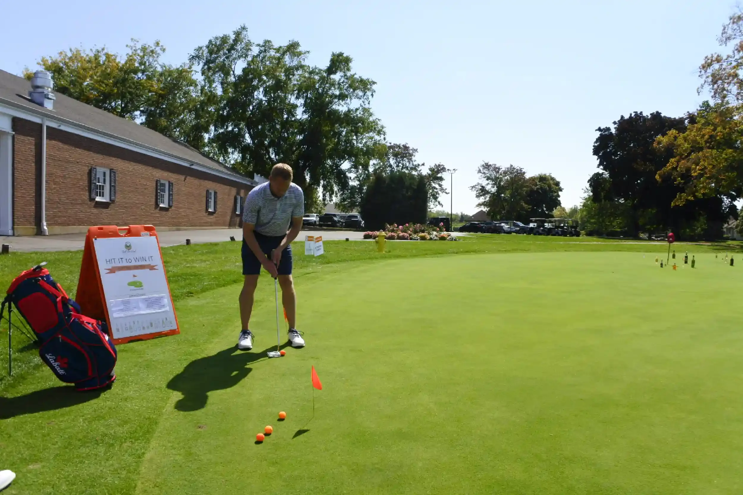 A man practicing golf on a putting green, preparing to hit the ball, with a golf bag, a sign, and golf balls nearby, on a sunny day.