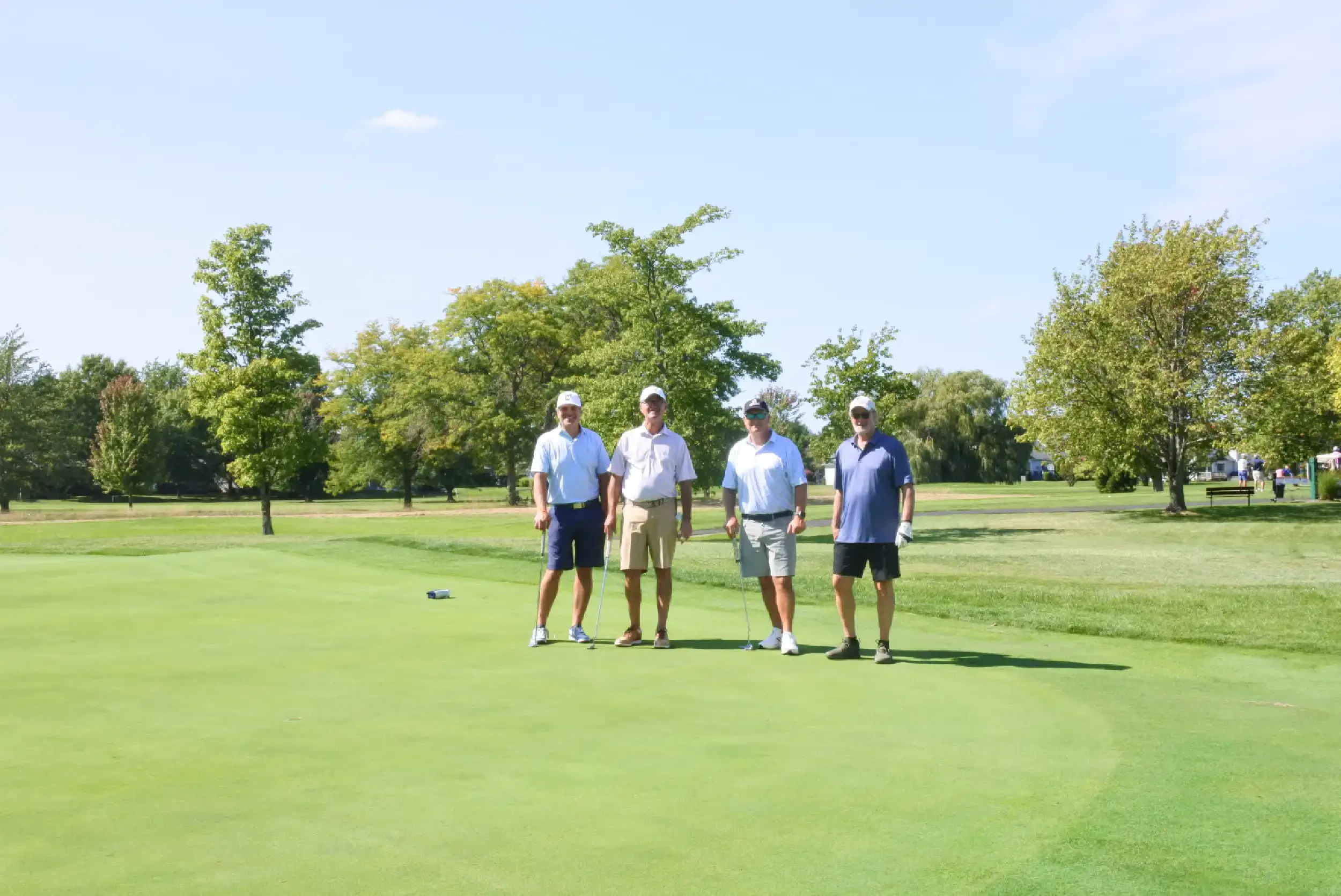 Four men standing on a golf course, holding golf clubs, with trees and a clear sky in the background.