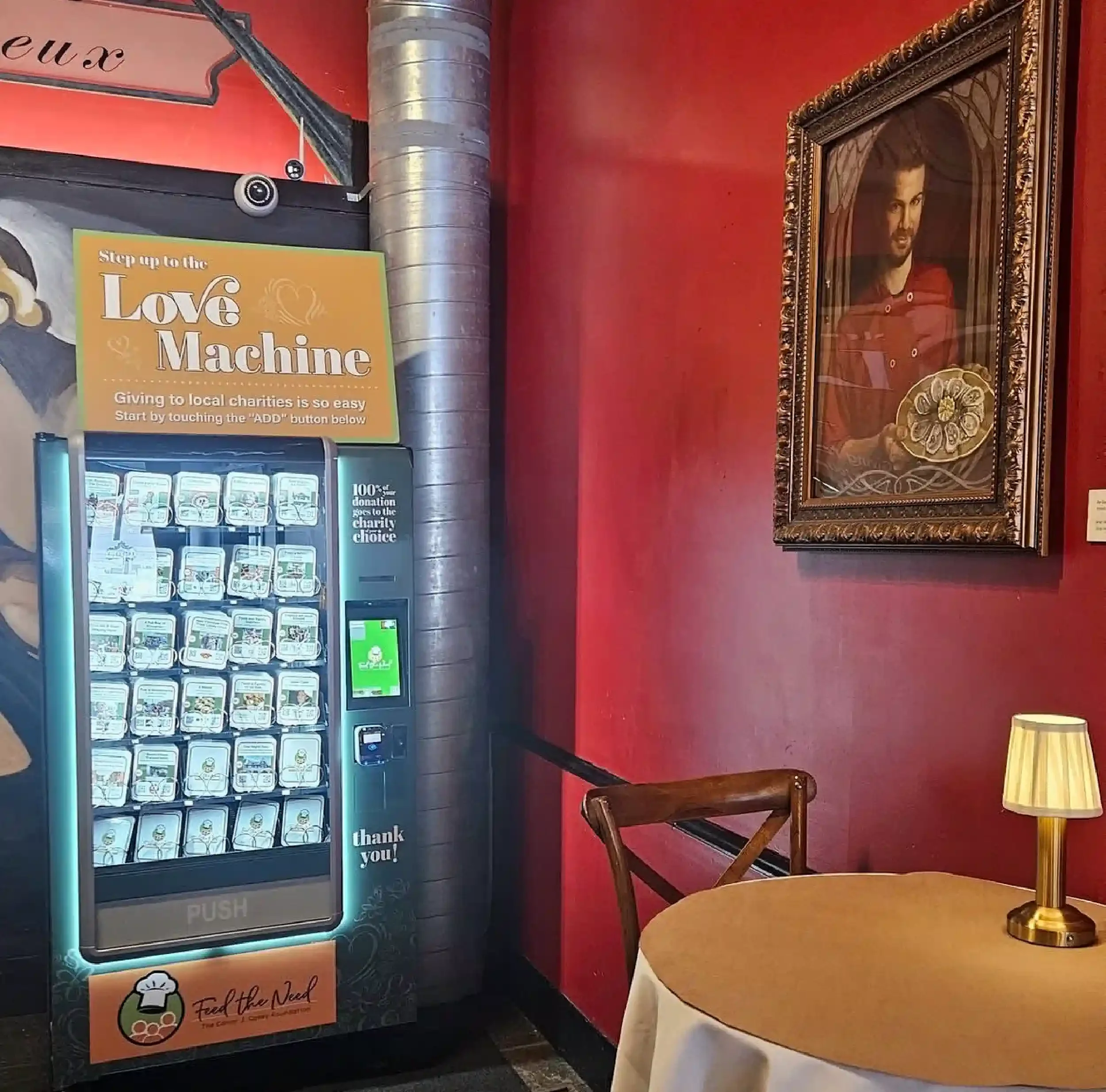 Vending machine with donation options for charity, a sign that says 'Stop up to the Love Machine,' and a framed portrait of a man on a red wall. There is a table with a gold-colored lamp in the foreground.