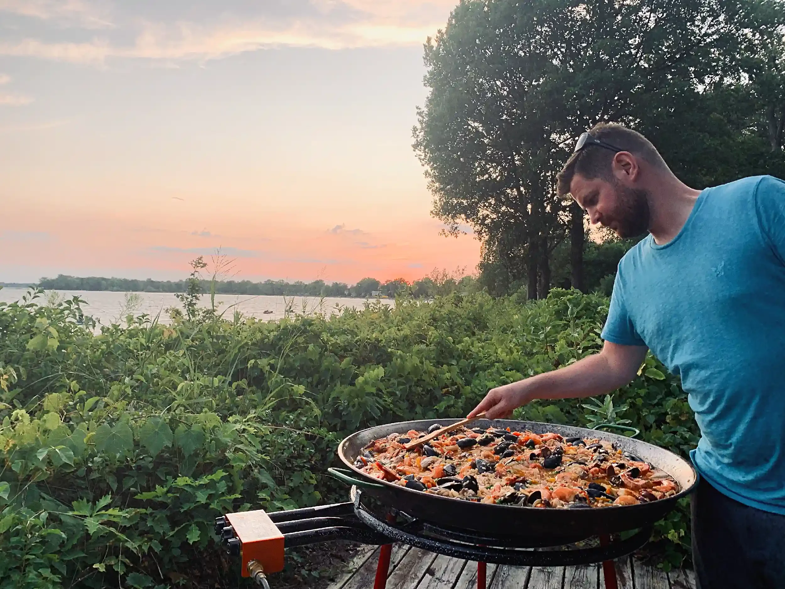 A man in a blue t-shirt cooking a large seafood paella outdoors at sunset near a river, with trees and a partly cloudy sky in the background.