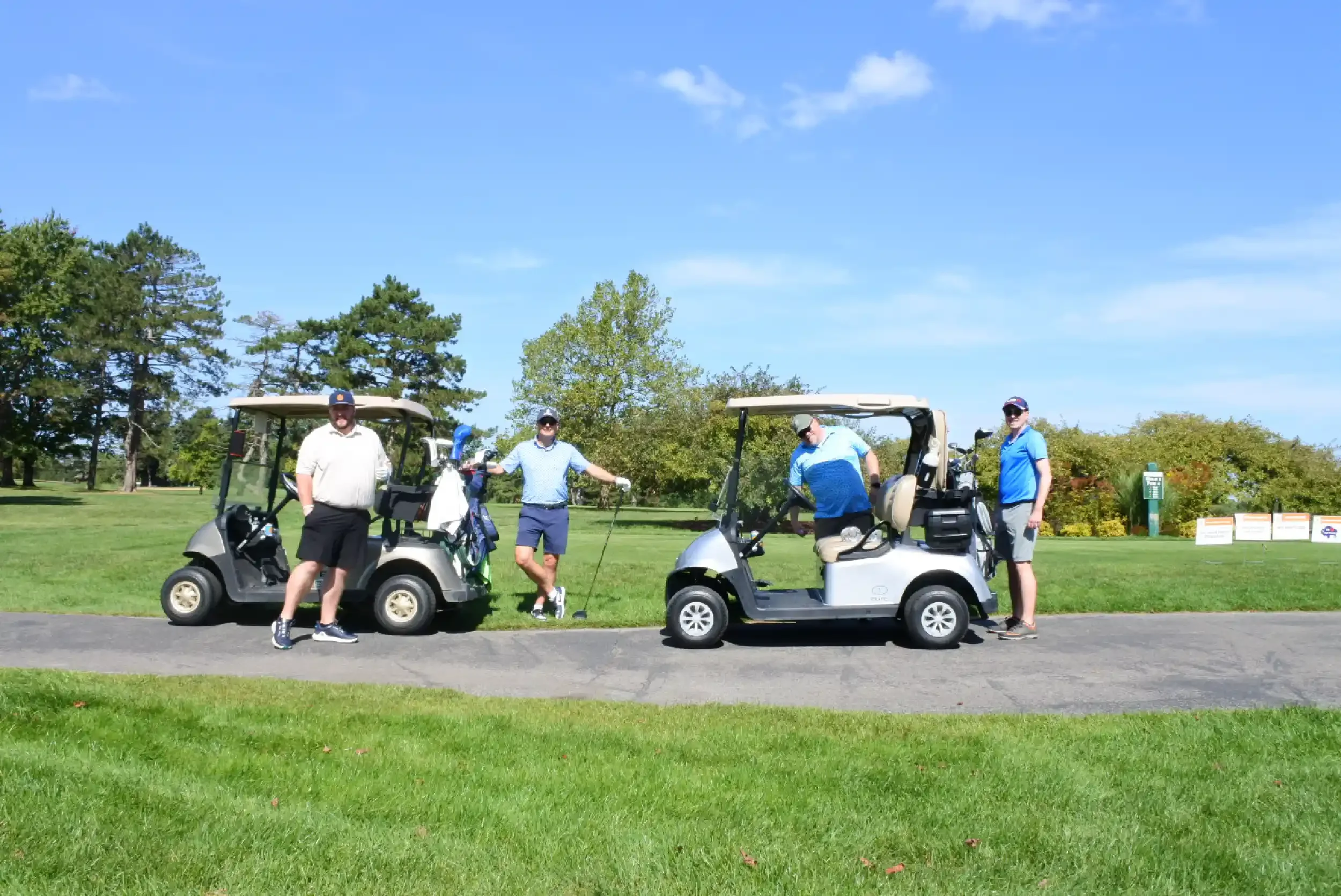 Four men standing next to two golf carts on a golf course during daytime. Two men are leaning against the golf carts, which are parked on a paved path surrounded by green grass and trees. The sky is blue with a few scattered clouds.