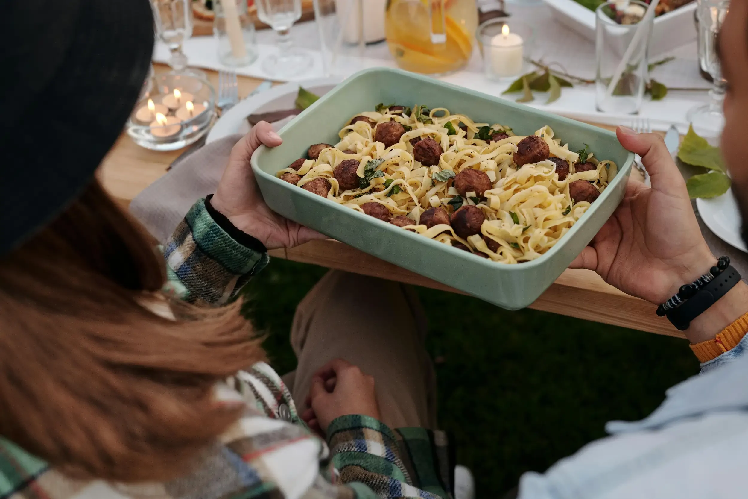 A person holding a large teal bowl of pasta with meatballs and greens at a table set for a meal, with other glasses, candles, and decorations visible in the background.