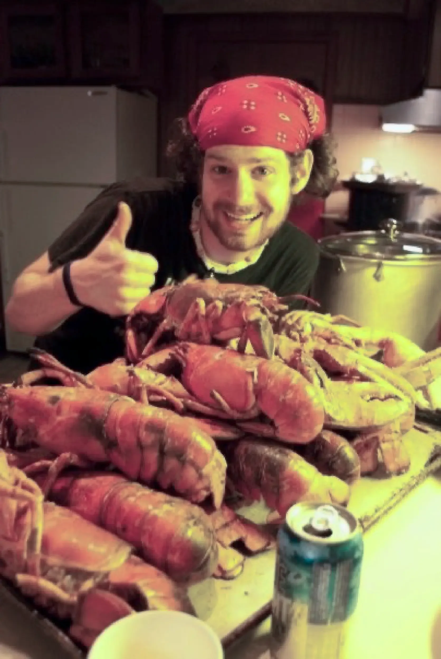 A man with curly hair wearing a red bandana is smiling, giving a thumbs-up, and sitting behind a large pile of cooked lobsters on a table with a yellow tablecloth in a kitchen.