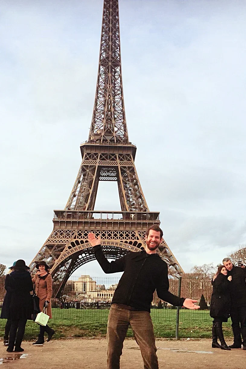 A man posing in front of the Eiffel Tower in Paris, France, with other visitors nearby on a cloudy day.
