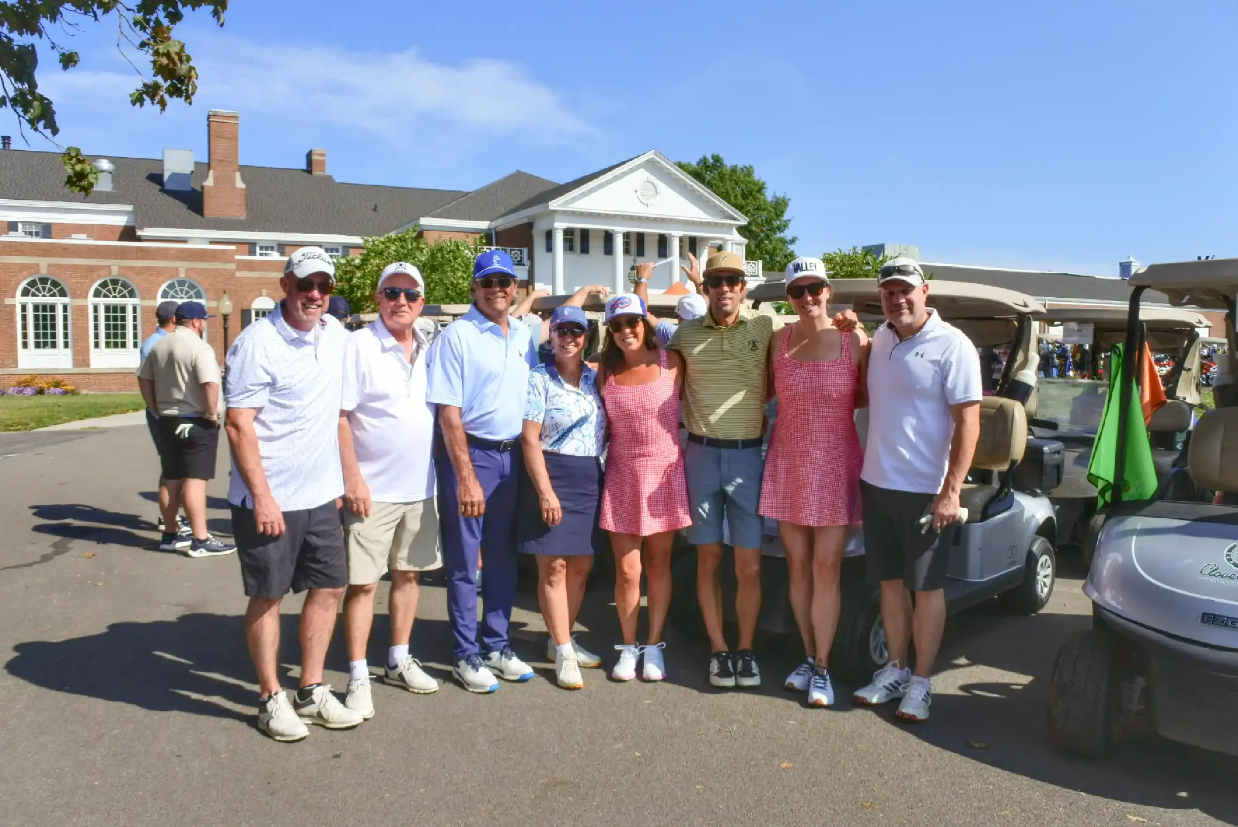 A group of nine people standing together outdoors on a sunny day, with a building and three golf carts in the background. The group includes men and women dressed in casual and golf attire, smiling and posing for the photo.