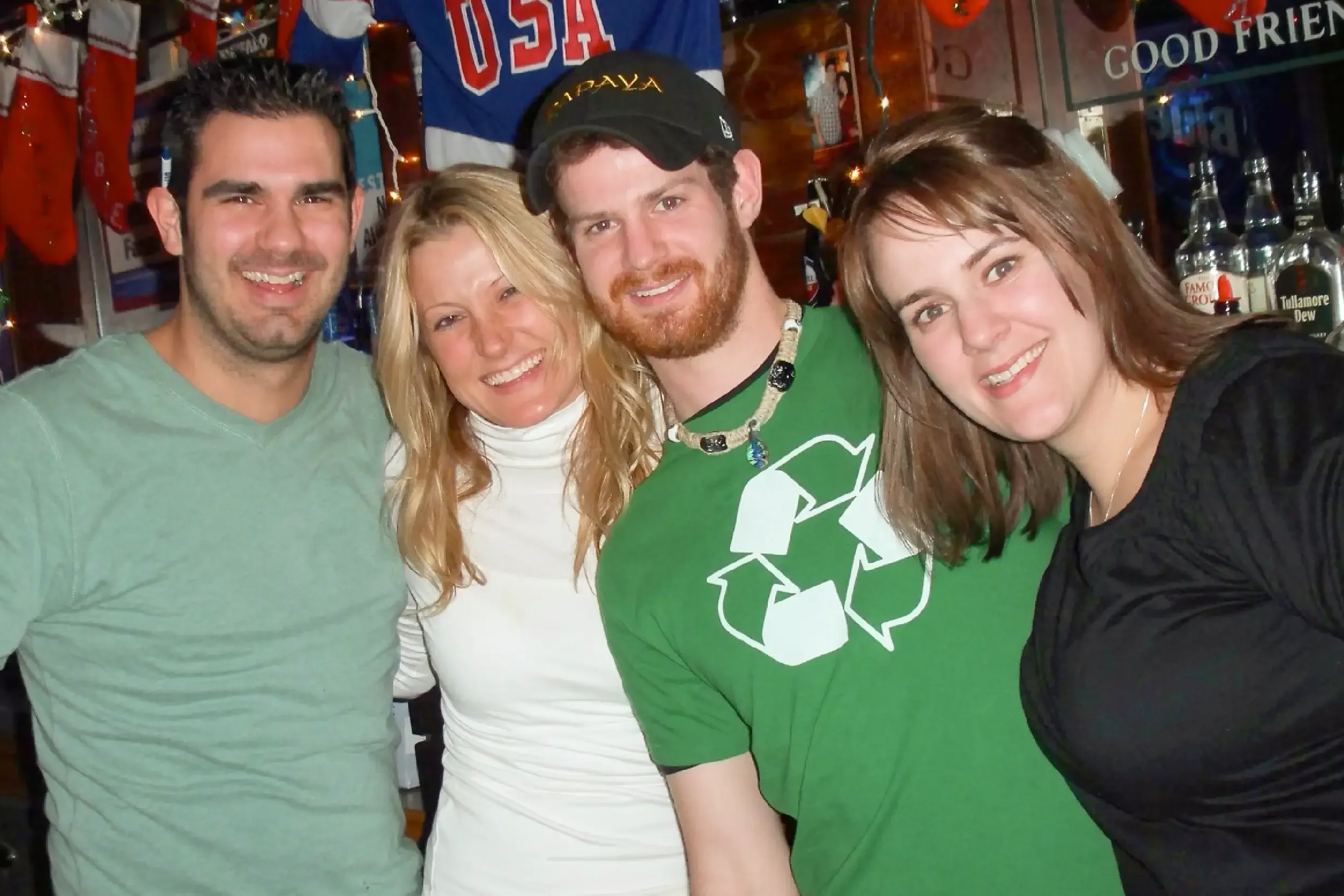A group of five people smiling and posing for a photo in a bar or pub decorated with sports memorabilia and Christmas stockings. The group includes three men and two women, dressed casually.