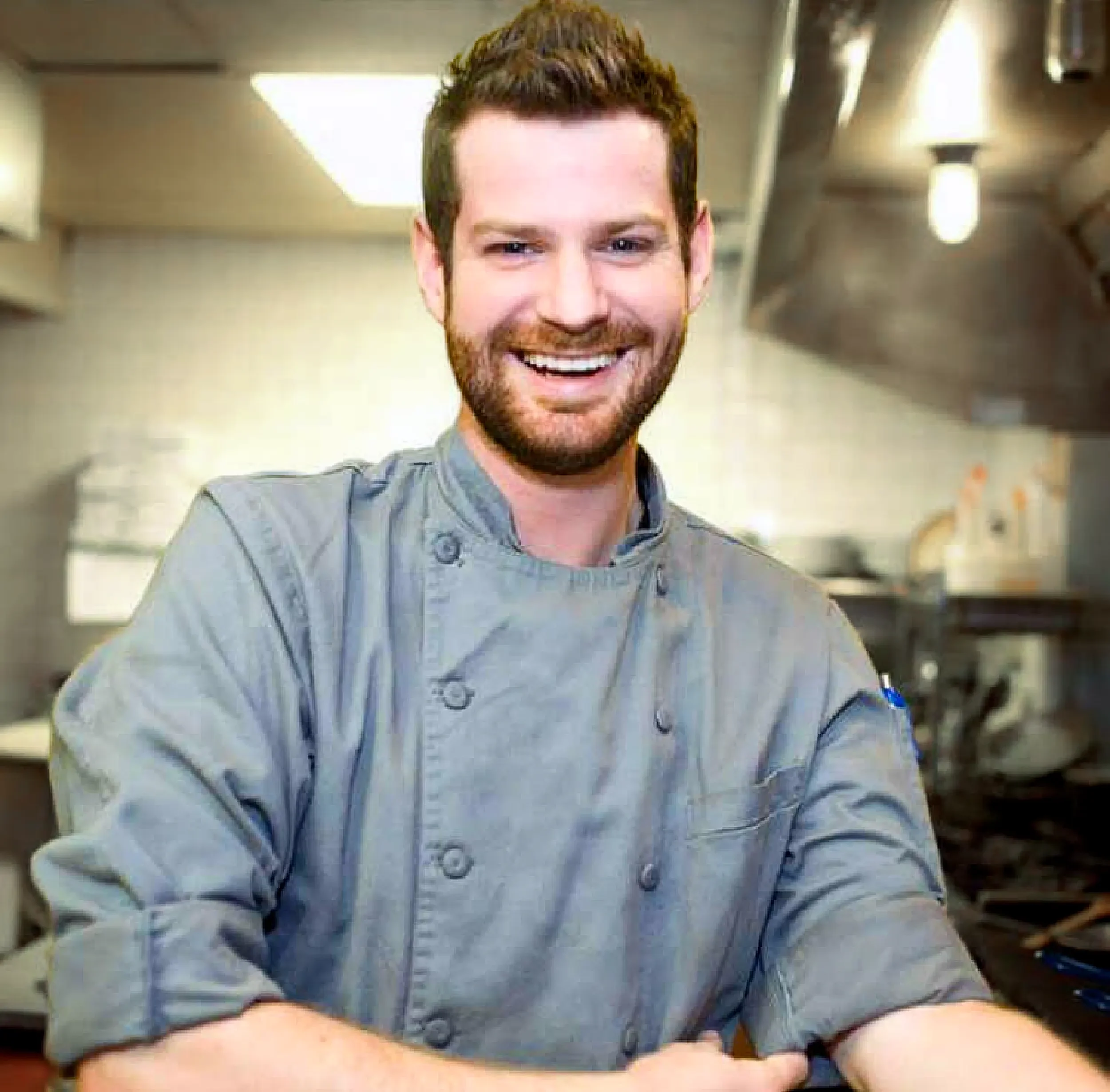 A smiling man with a beard and short brown hair, wearing a blue-gray button-up shirt, is seated at a table in a kitchen or dining area.