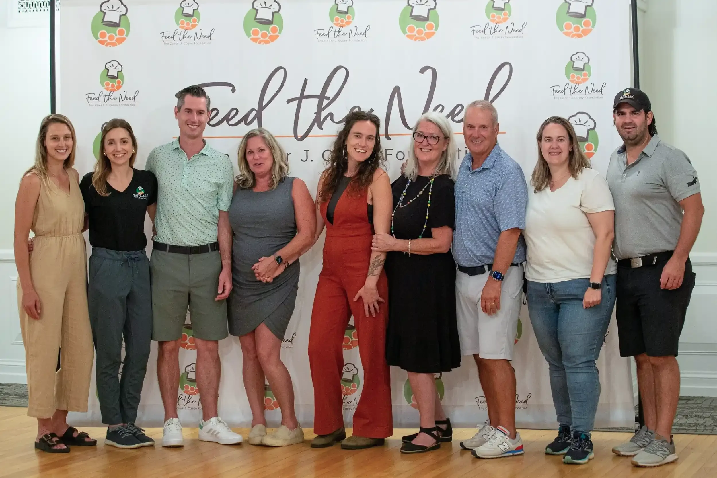 Group of nine people standing in front of a backdrop with the 'Feed the Need' logo, smiling.