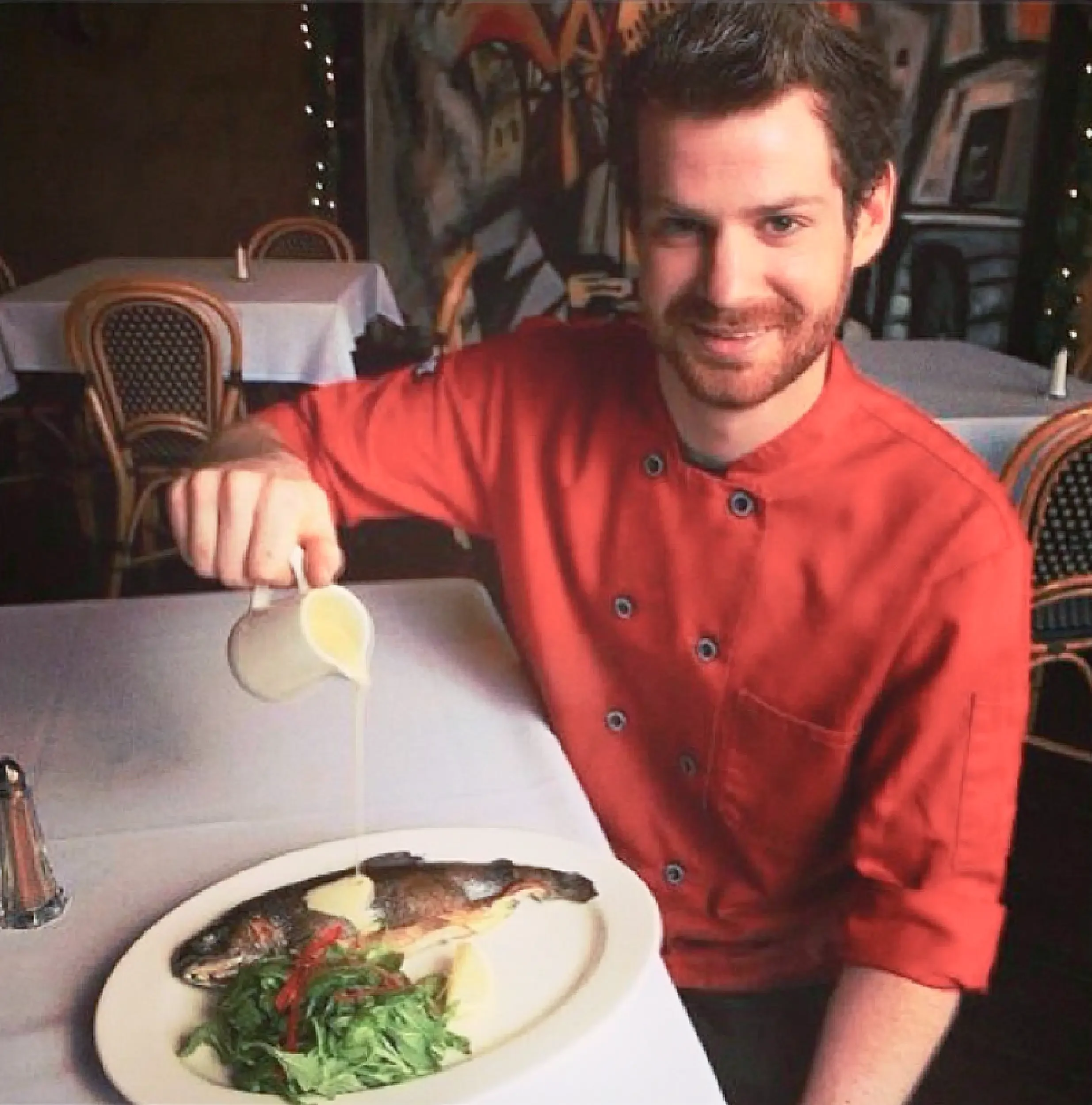 A man with dark hair and a beard, wearing a red shirt, is smiling at the camera, taking a selfie in a restaurant. On the table in front of him, there is a bowl of salad with greens, tomatoes, and other vegetables. The restaurant has decorative elements on the wall and a table with a white tablecloth and chairs with a woven pattern.