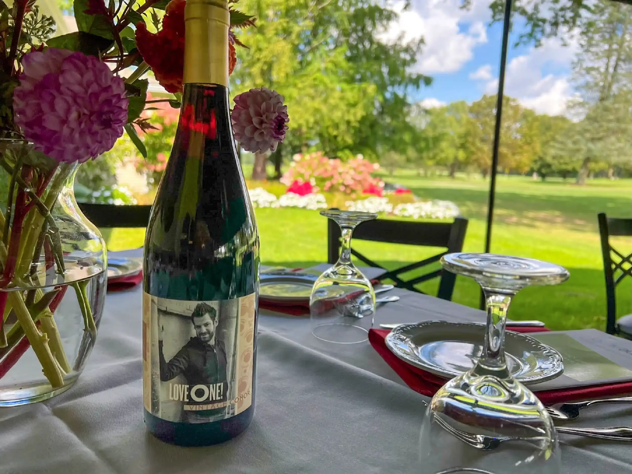 A bottle of wine with a vintage-style label featuring a man and the text 'LOVE ONE!' on a table set for an outdoor dining area, with glasses, plates, and floral arrangements, with lush green trees and blue sky in the background.