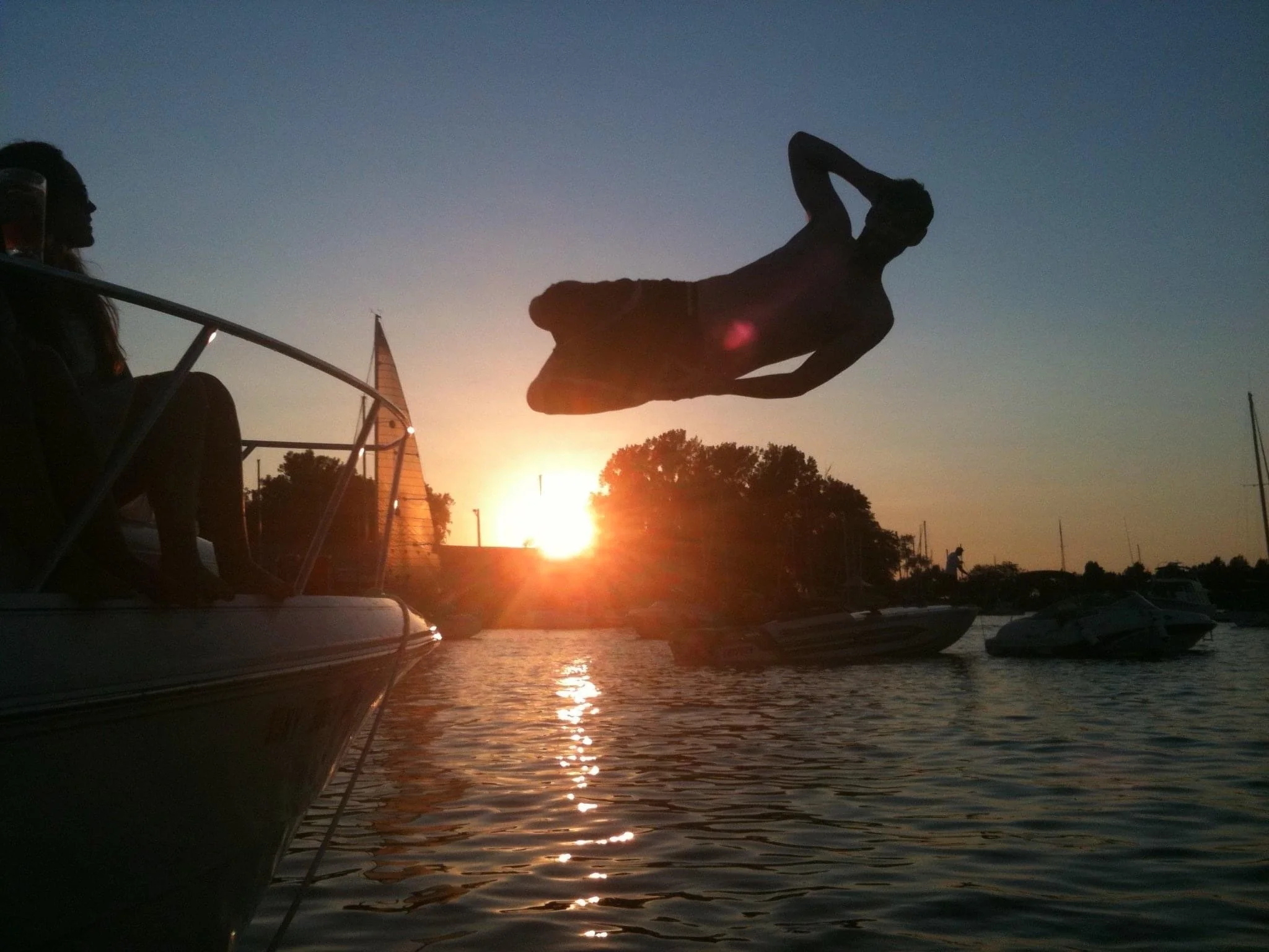 A silhouetted figure performs a “leisure dive” from a boat into a calm lake at sunset, with golden light reflecting across the water and other boats nearby, capturing a relaxed summer evening on the water.