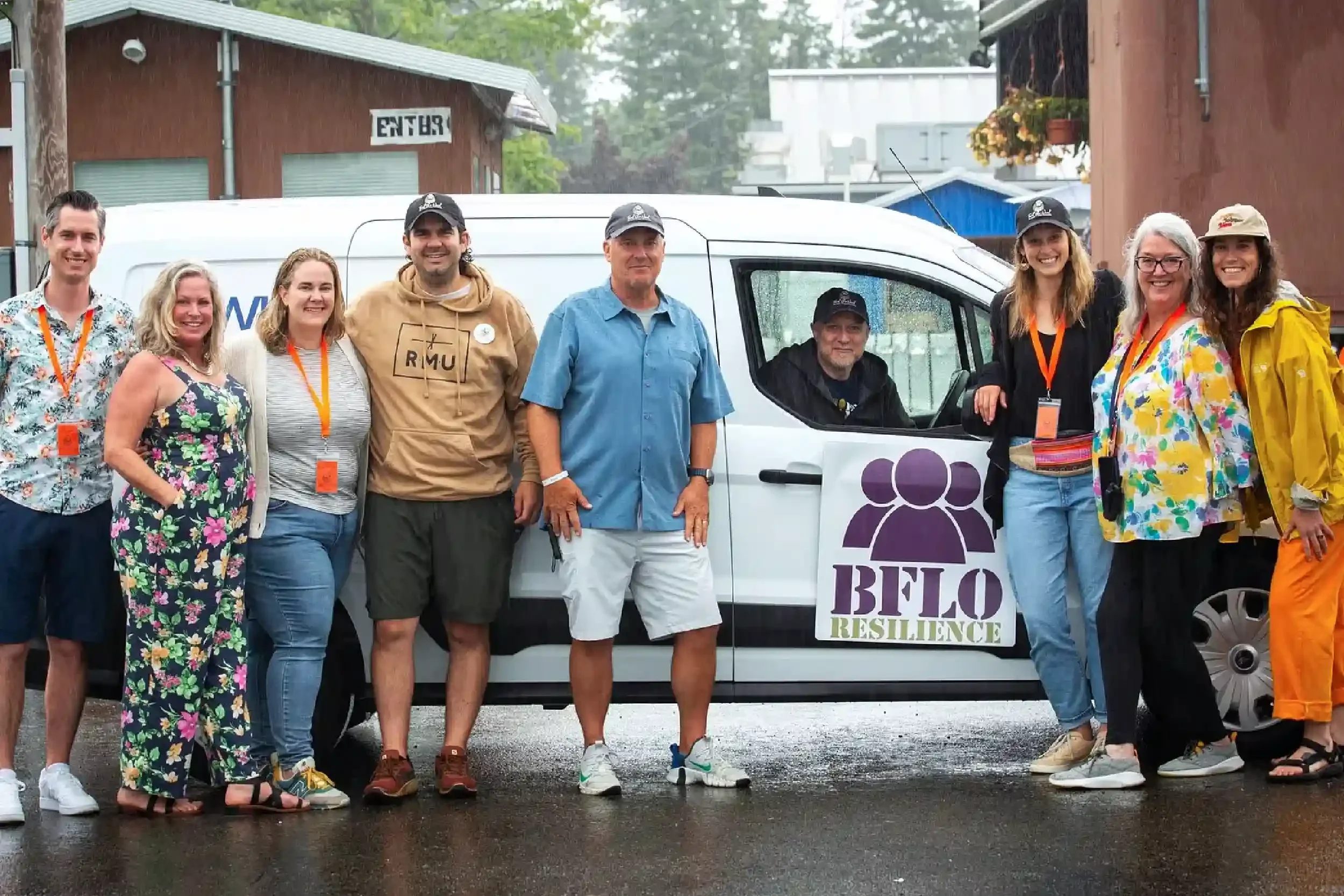 Group of nine people standing outdoors in front of a white van with a purple logo and a sign that reads "BELO RESILIENCE". It is raining, and the group is smiling, dressed in casual clothes, some with rain jackets and lanyards.