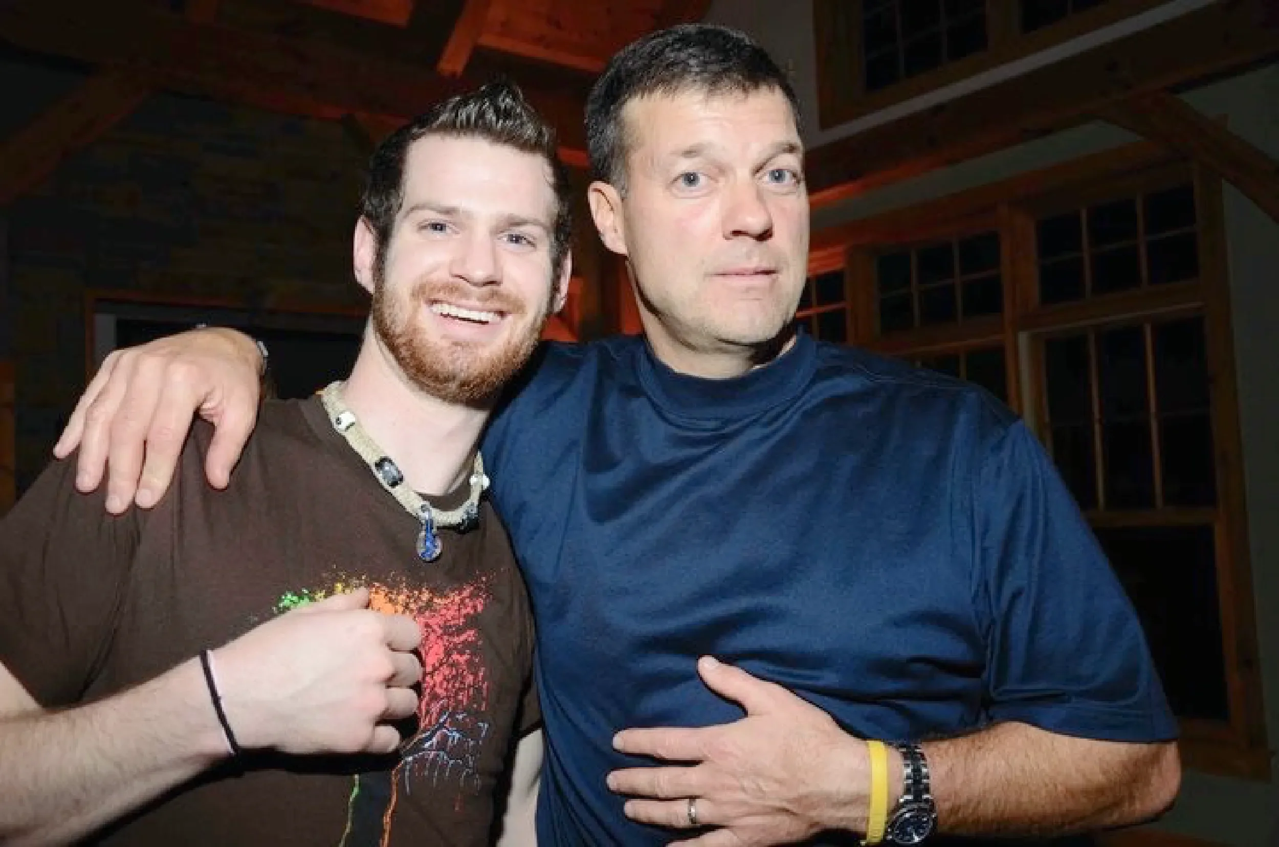 Two men taking a selfie. The man on the left has a beard and is smiling, wearing a brown shirt and beaded necklace. The man on the right is looking directly at the camera, wearing a blue shirt and a wristwatch. They are in a room with a wooden ceiling and shelves in the background.