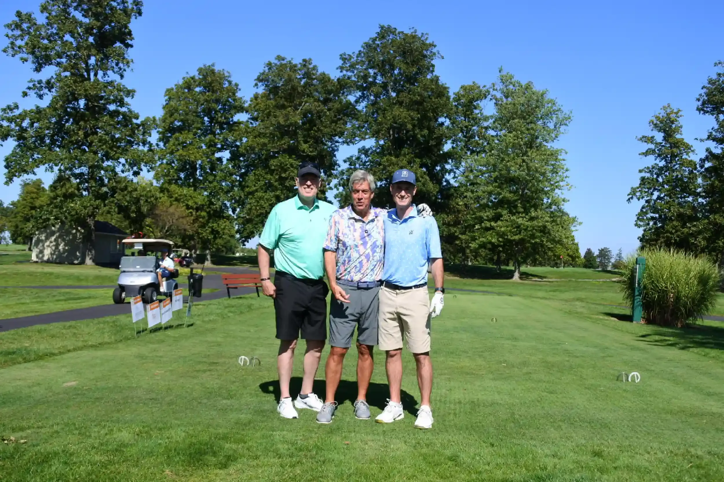 Three men standing on a golf course with trees and a small building in the background, smiling and posing for the photo.