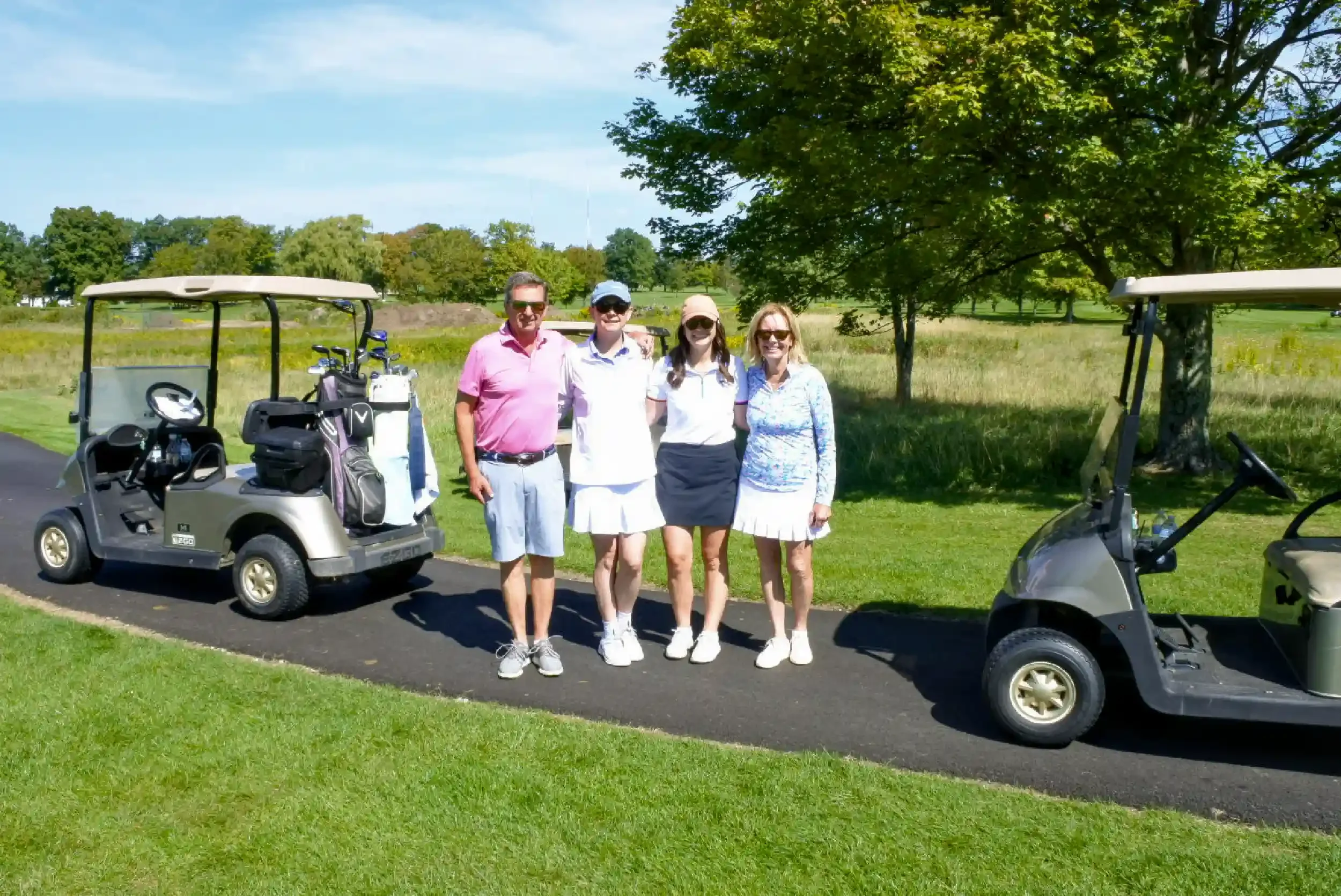 Four people standing together on a golf course path with two golf carts nearby, trees and blue sky in the background.
