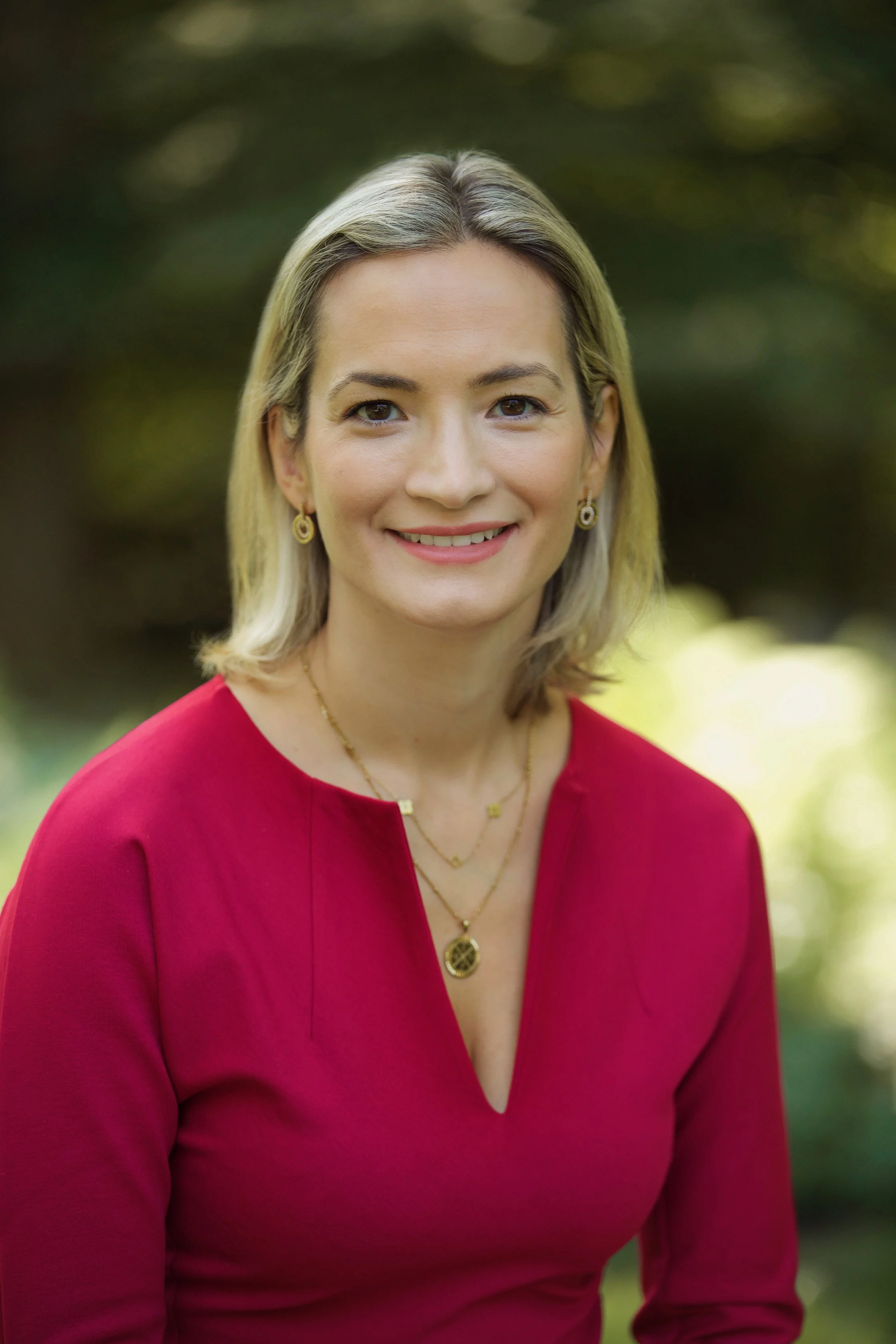 A woman with blonde hair smiling outdoors, wearing a red dress and gold jewelry, with a blurred green background.