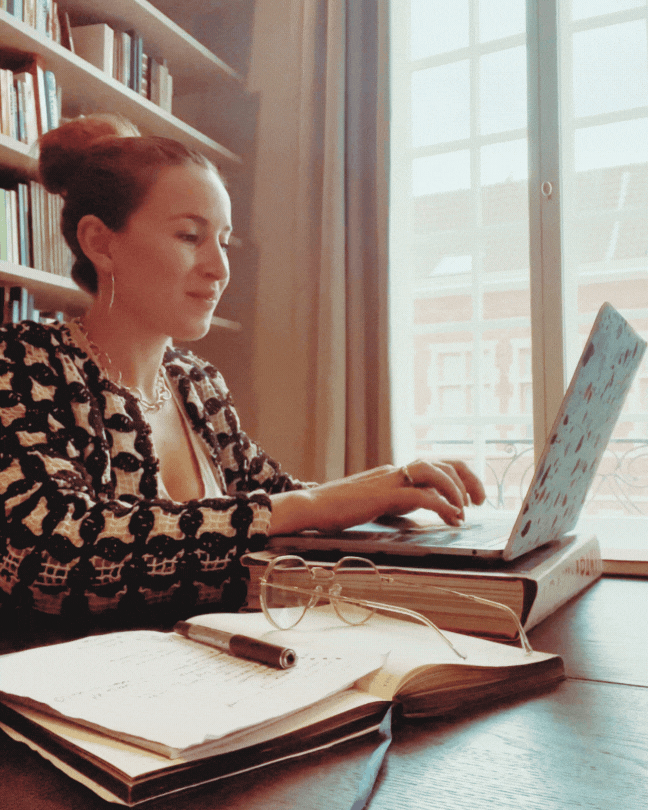 A woman sitting at a table with a laptop, open notebook, glasses, and a pen, in a bright room with a window and bookshelf.