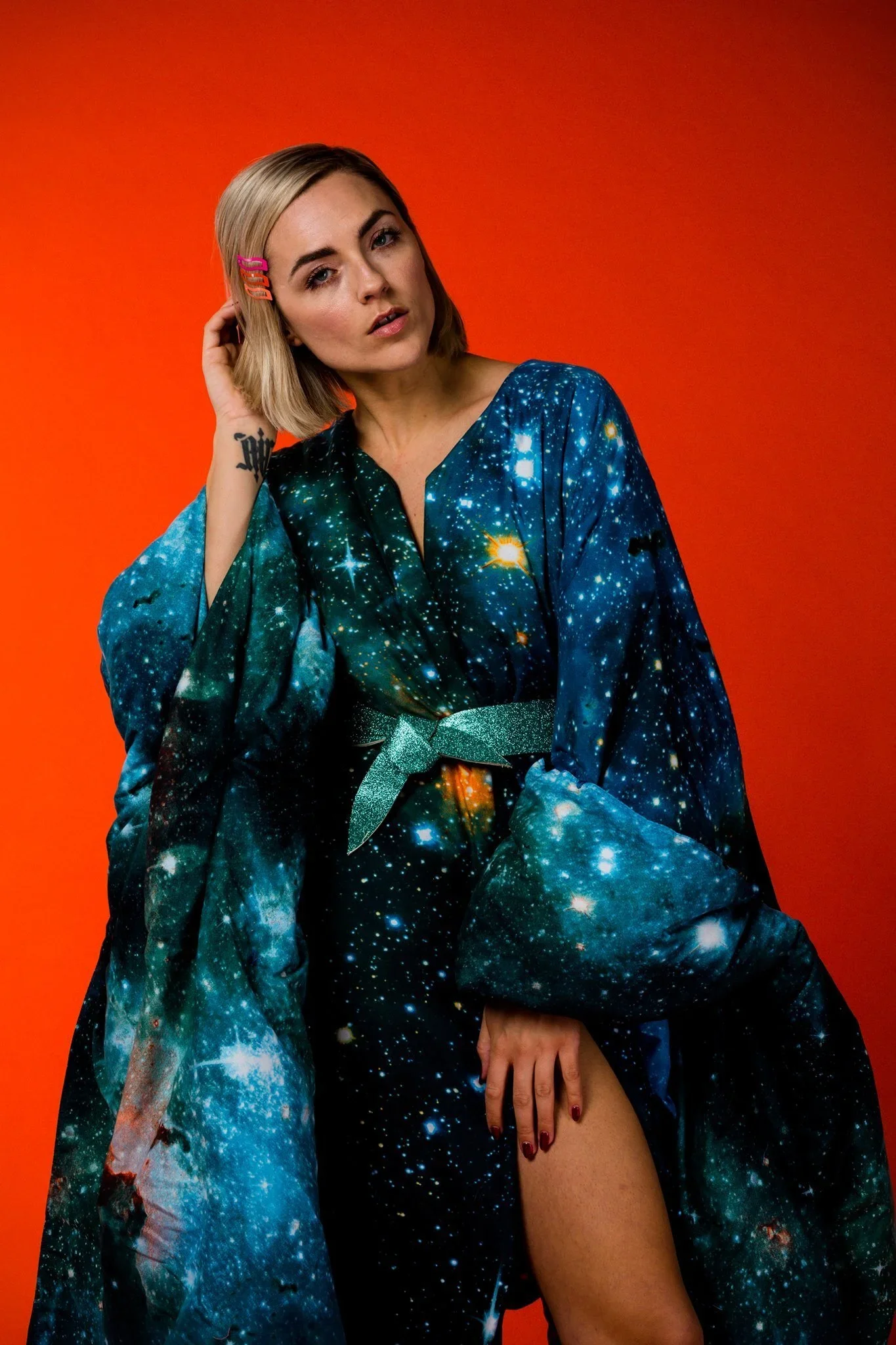 A woman with blonde hair wearing a galaxy print robe and a glittery teal belt, posing against an orange background.