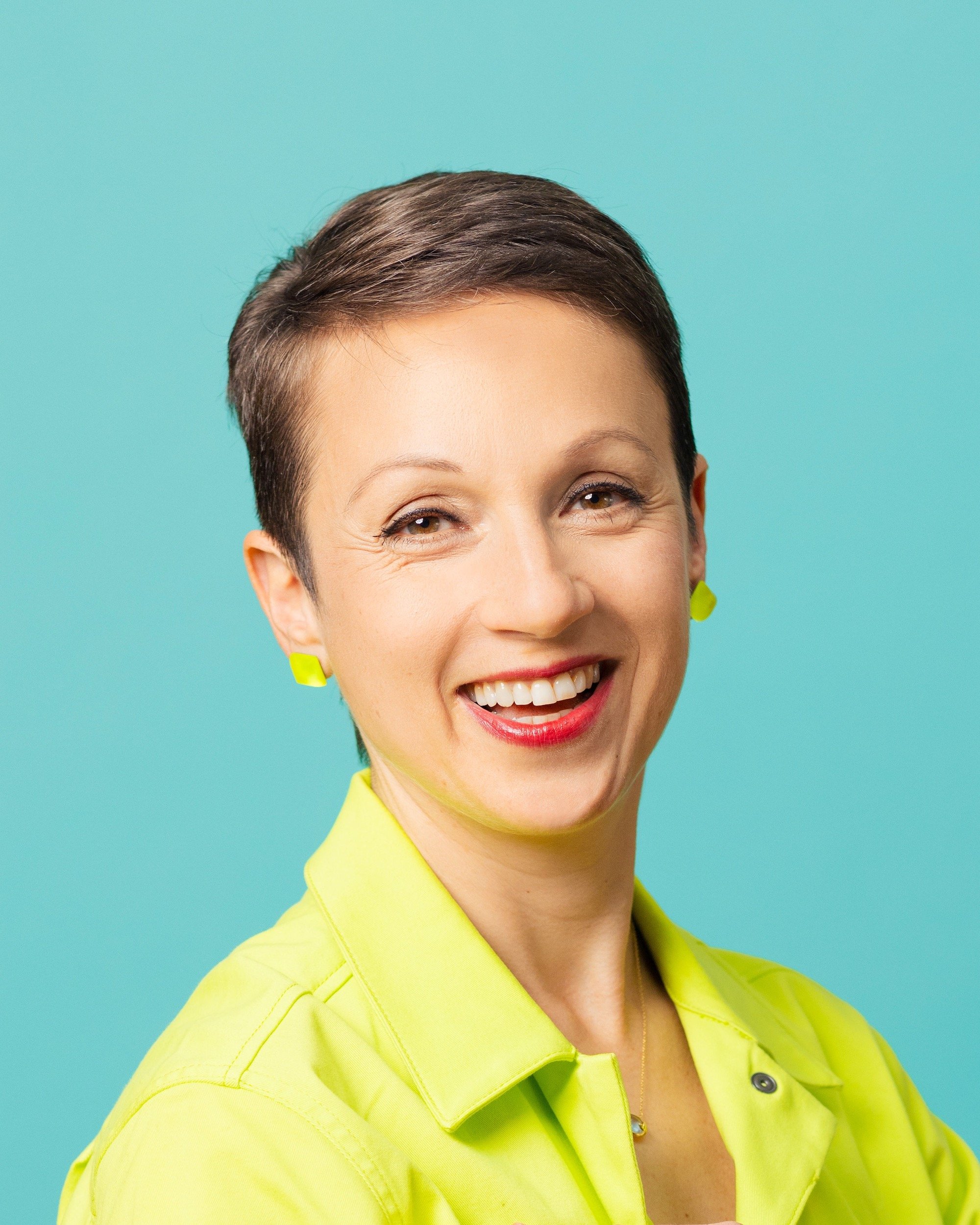 A woman with short dark hair, smiling, wearing a bright yellow shirt and matching earrings, with a solid turquoise background.