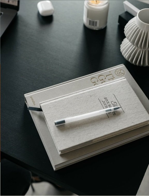 A black desk with a closed book titled "The Tailor of Gloucester," a white pen resting on top, a white candle, a small white container, a tissue box, and a ribbed, circular white decorative item.