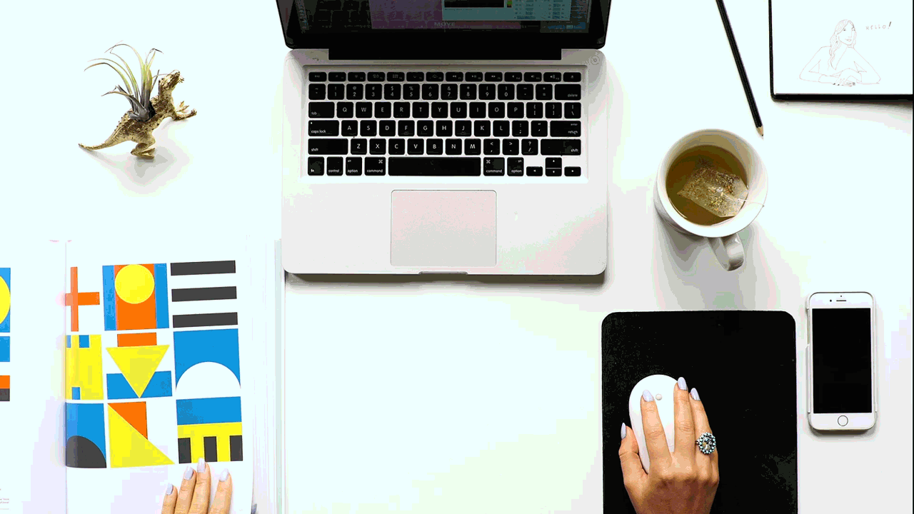 Top-down view of a workspace with a laptop, a tablet with sketches, a cup of tea, a smartphone, a mouse on a mouse pad, a colorful notebook, and a small decorative dinosaur plant.