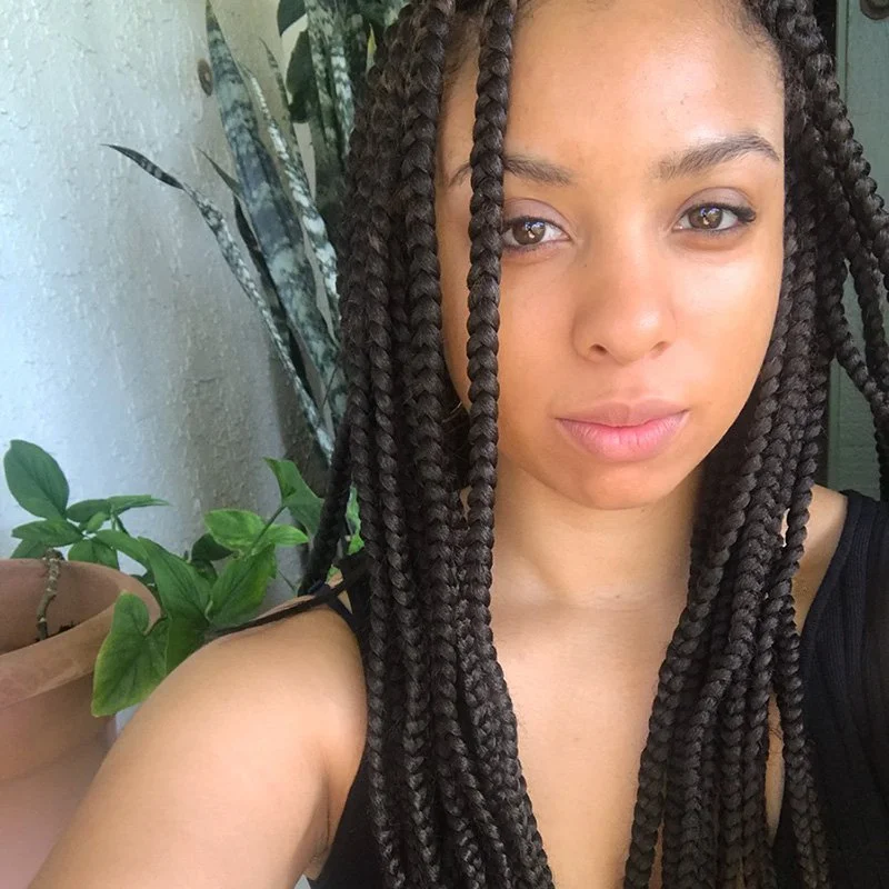 Close-up of a young woman with long box braids, wearing a black top, standing outdoors near green plants and a textured white wall.