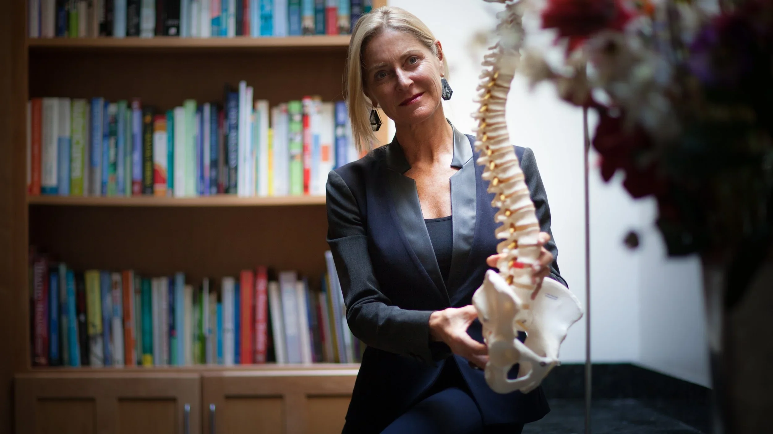 A woman with blonde hair in a black blazer showing a human spine model in a room with a bookshelf filled with books.
