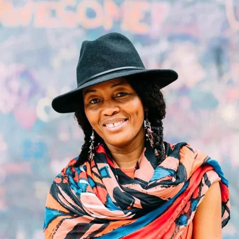 A woman smiling, wearing a black hat, colorful scarf, and earrings, with a blurred background.