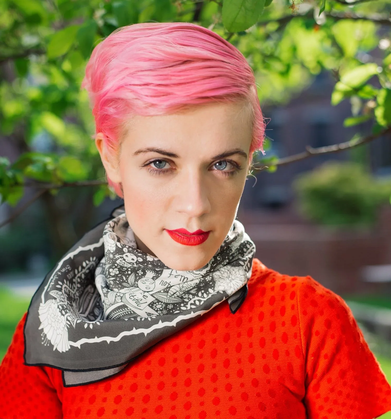 Woman with pink hair, red lipstick, and light blue eyes, wearing a black and white patterned scarf and a red textured top, outdoors with green leaves in the background.