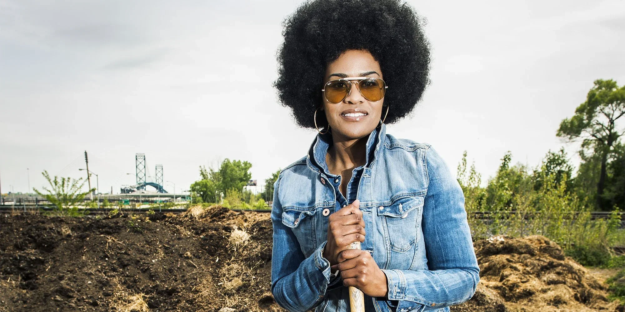 Woman with curly afro hairstyle wearing sunglasses, hoop earrings, and a denim jacket holding a shovel outdoors with trees and a bridge in the background.