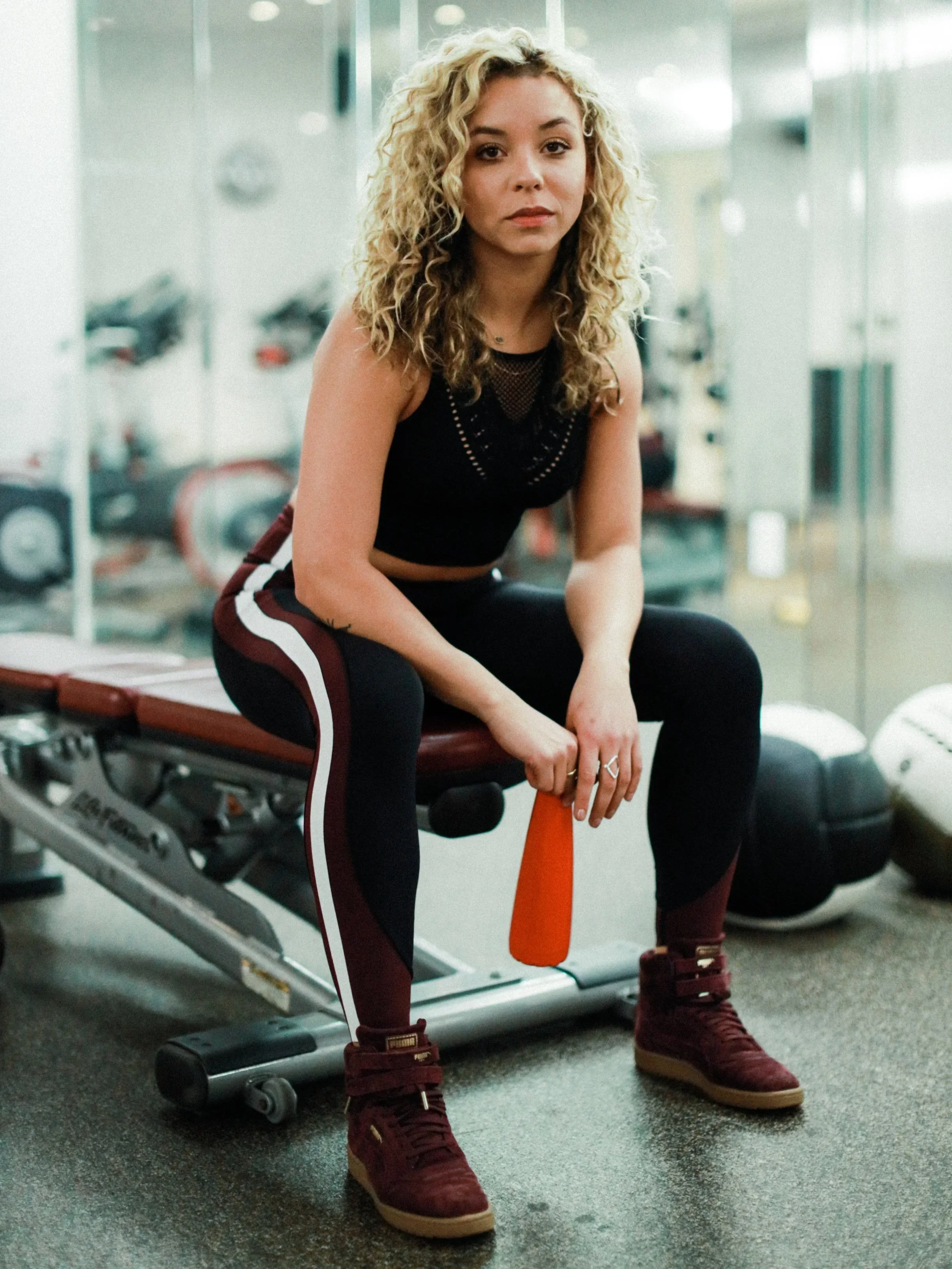 A young woman with curly blonde hair sitting on a workout bench in a gym, holding an orange water bottle, wearing a black sleeveless top, black leggings with white and burgundy stripes, and burgundy sneakers.