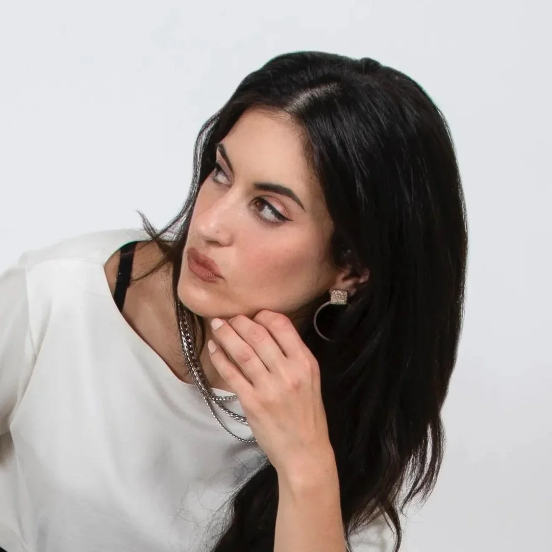 A woman with long dark hair wearing a white top and jewelry, resting her chin on her hand and looking to the side against a plain background.