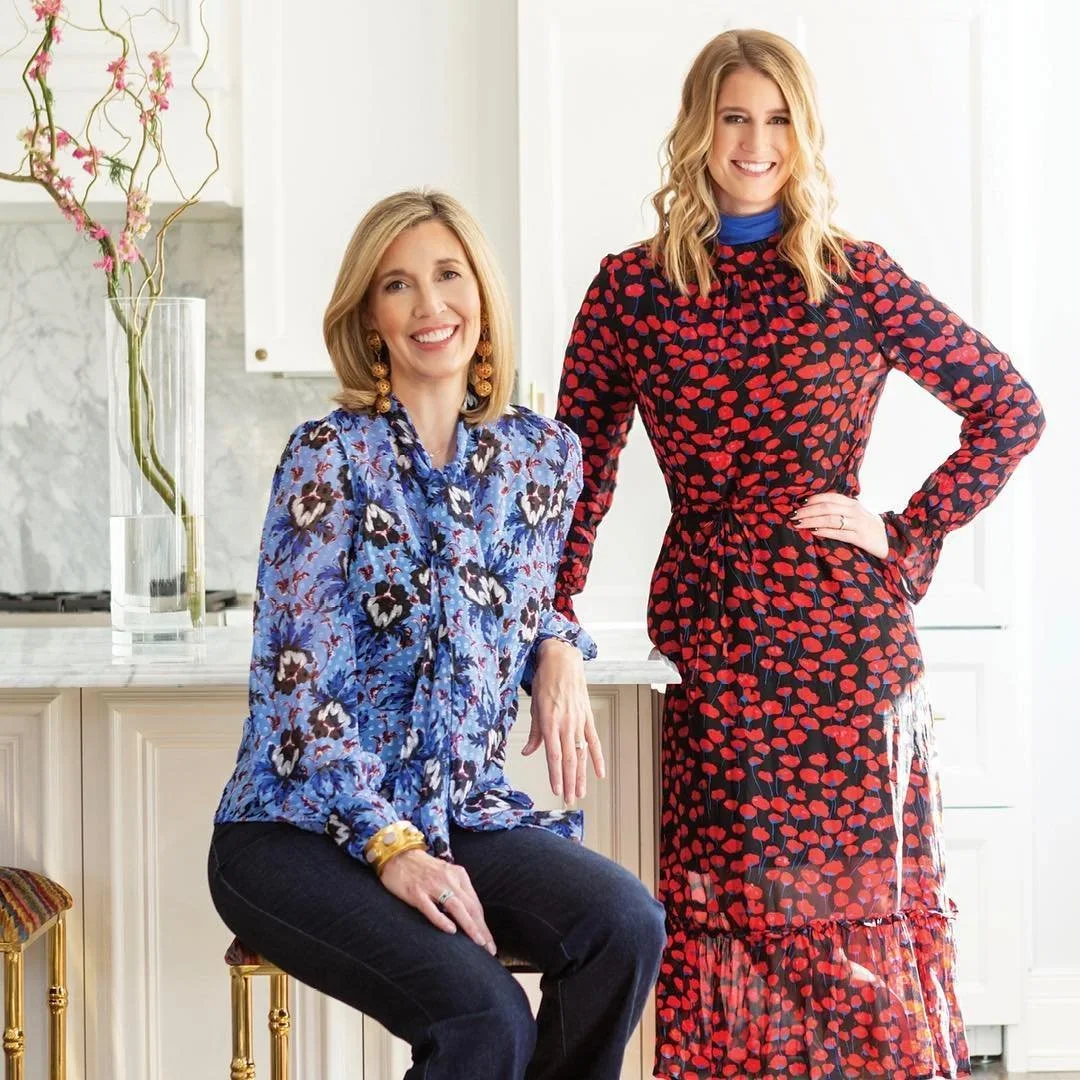 Two women posing in a bright kitchen, one sitting on a chair and the other standing, both smiling. One woman is wearing a blue floral blouse and black pants, and the other is in a red and black patterned dress.