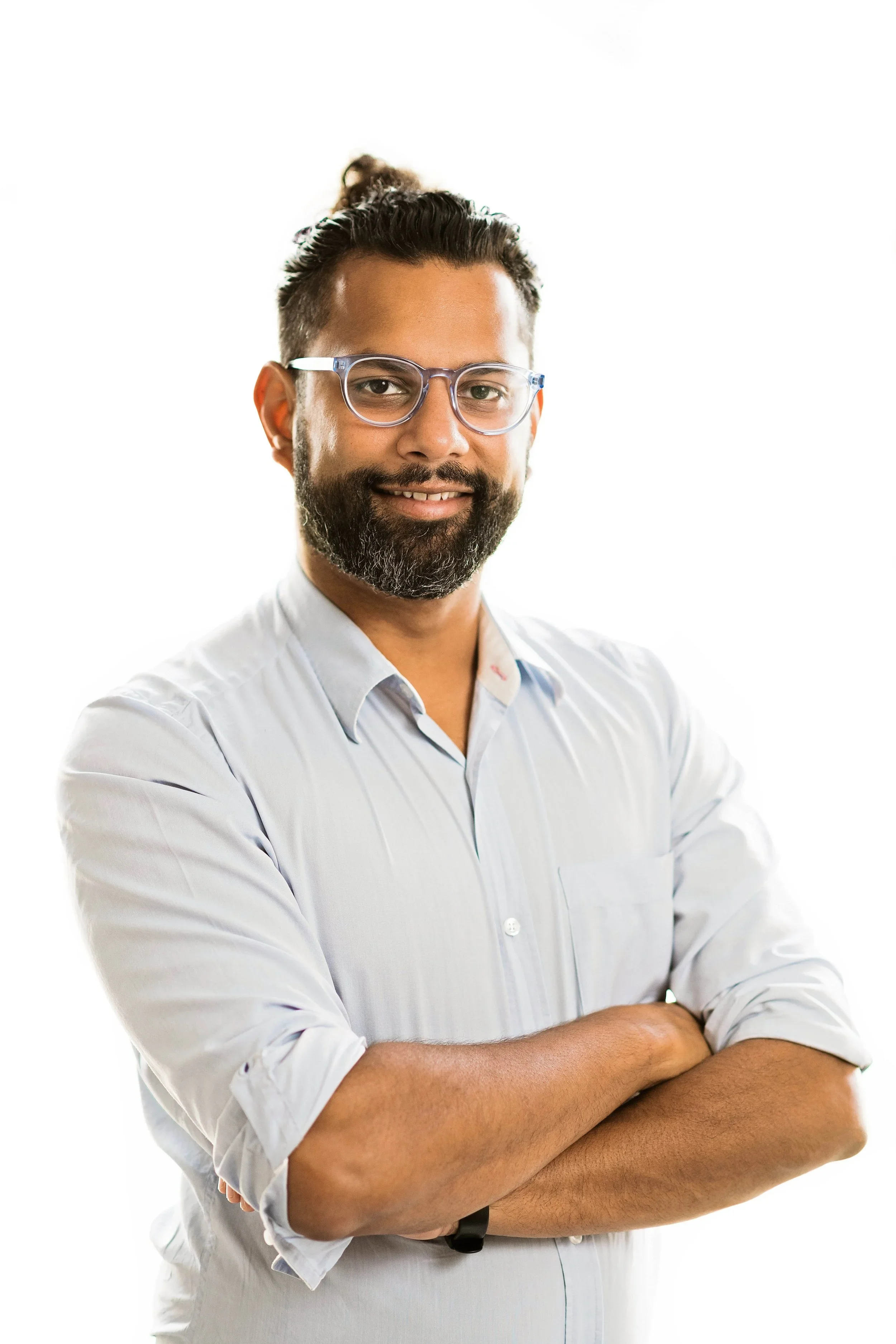 A bearded man with glasses, wearing a light blue button-up shirt, standing with arms crossed and smiling at the camera.