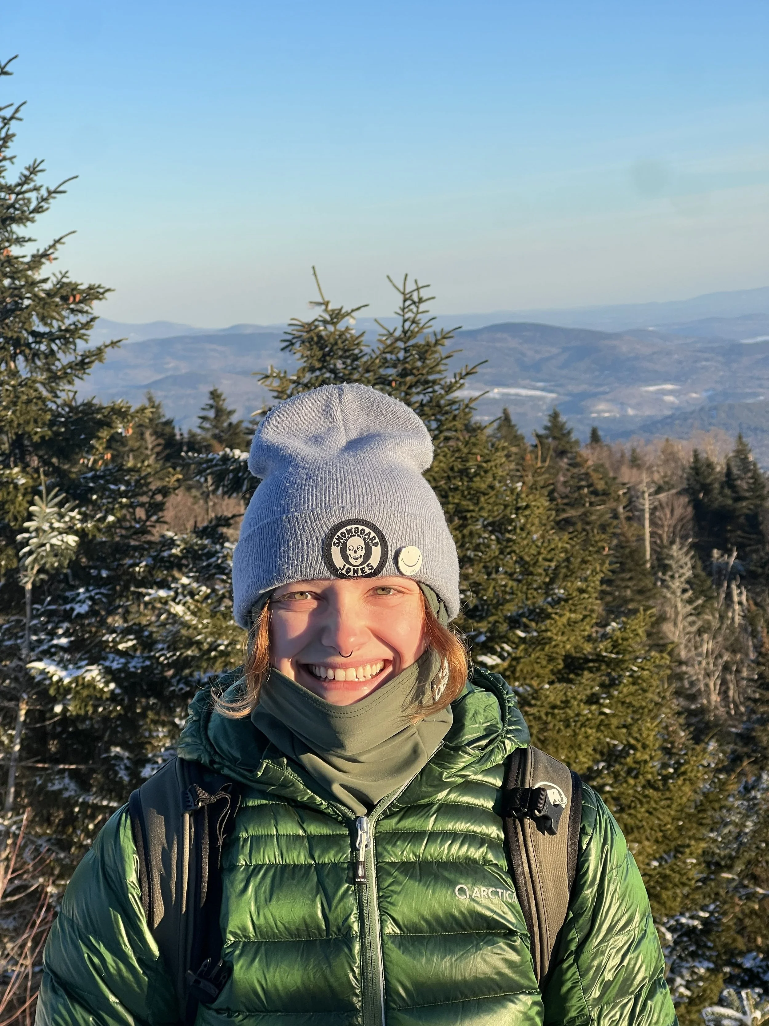 A smiling woman outdoors in a green puffy jacket and gray beanie with patches, surrounded by snow-covered evergreen trees and mountain scenery in the background.