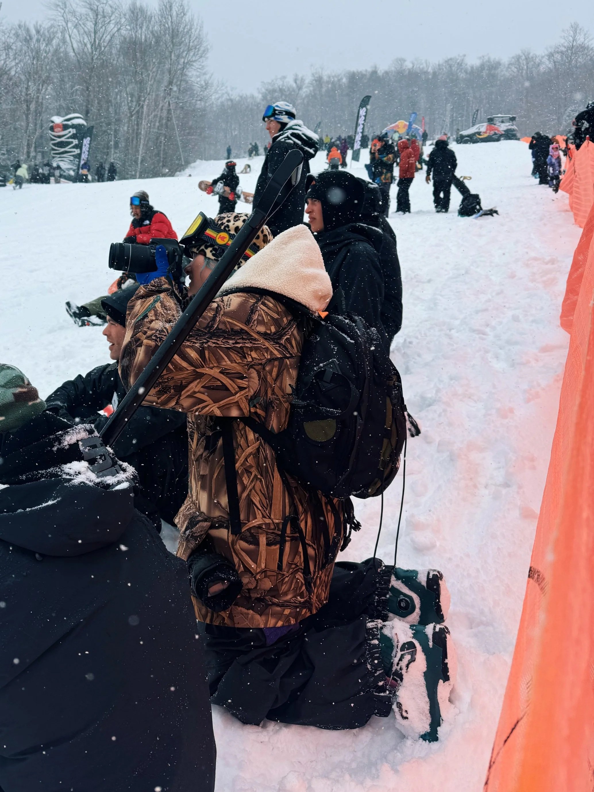 Group of people kneeling and sitting in snow at a winter event, surrounded by snow-covered trees, with some holding skis, snowboards, and cameras, and winter gear like jackets, helmets, and backpacks.