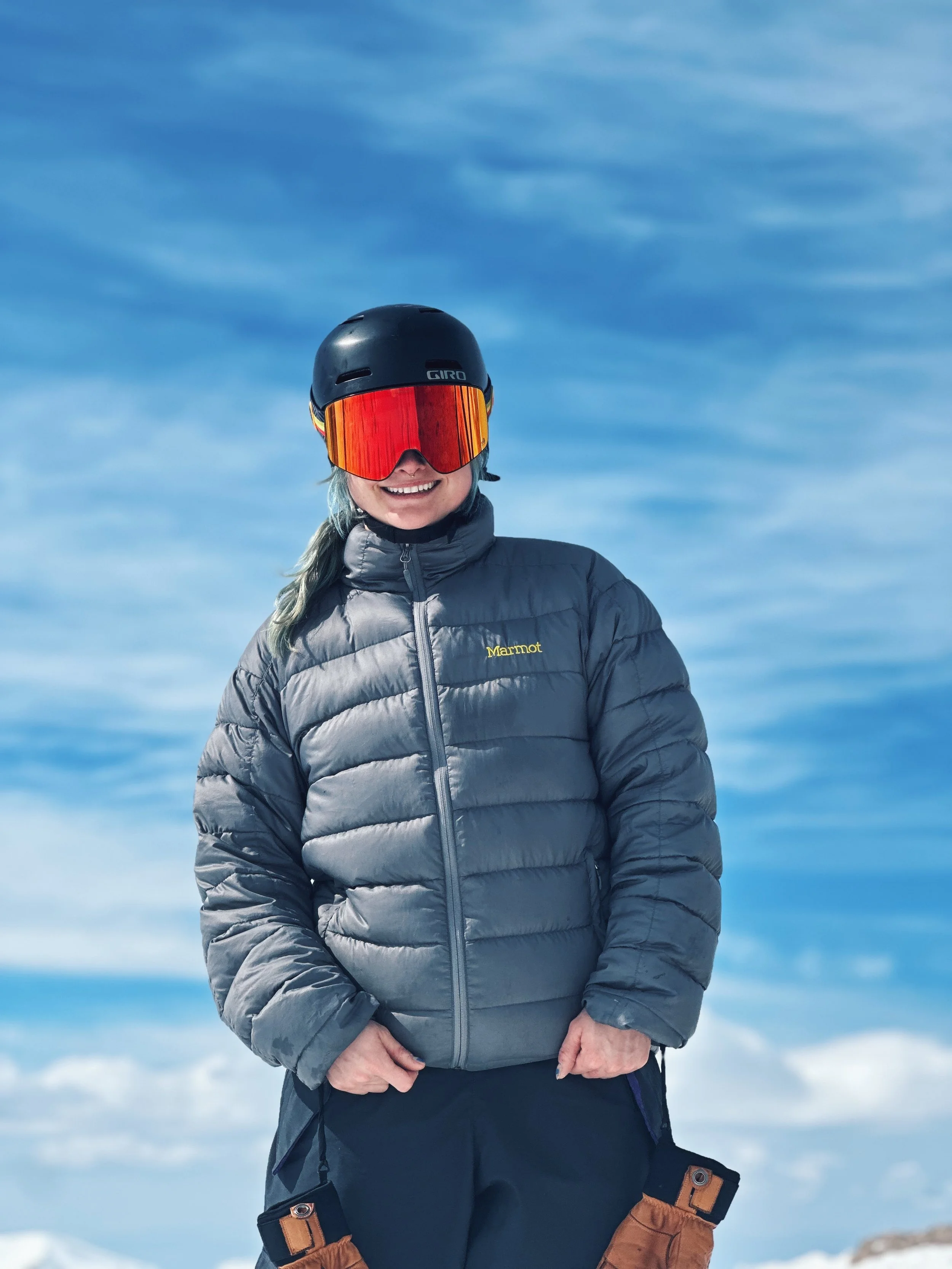 A woman dressed in winter sports gear standing outdoors against a snowy landscape and blue sky, wearing a helmet, ski goggles, and a puffy gray jacket.