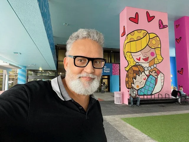 A man with gray hair and beard wearing glasses and a black shirt taking a selfie outside a colorful building with large pink cartoon mural of a woman hugging a child, with additional pink and purple walls and some people sitting near the mural.