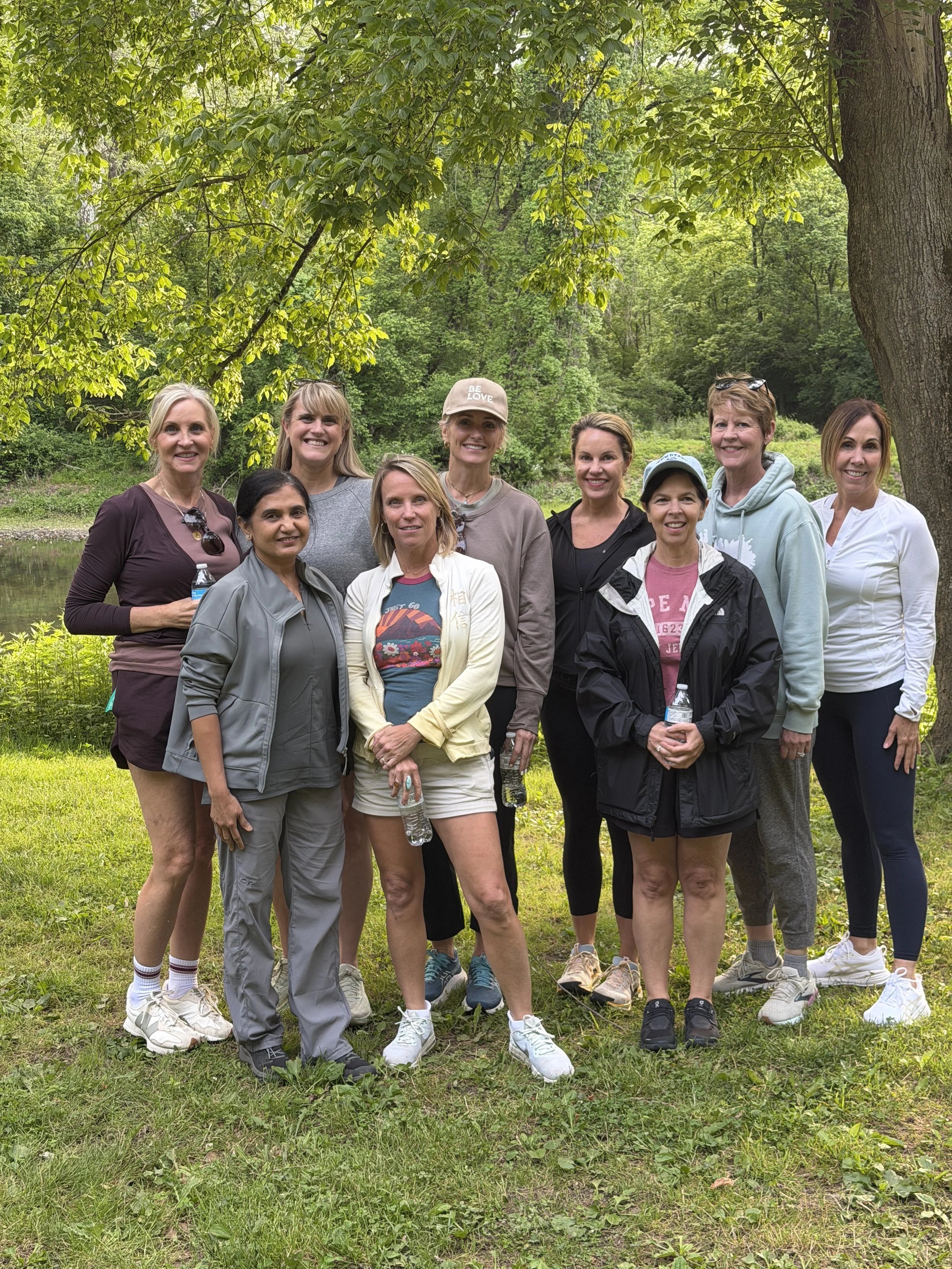 A group of women pausing together outdoors during a Trielle Collective walk, surrounded by trees and greenery near a creek