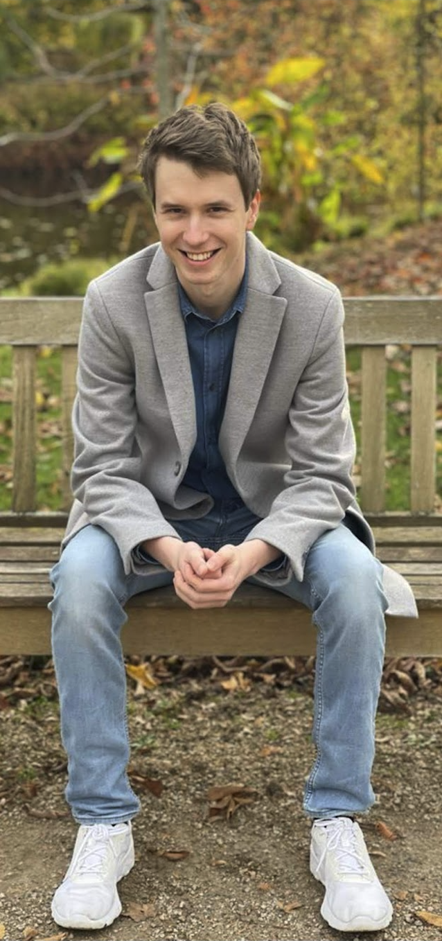A young man with dark hair, smiling, sitting on a wooden park bench during autumn, wearing a gray coat, blue shirt, light blue jeans, and white sneakers, with colorful fall foliage in the background.
