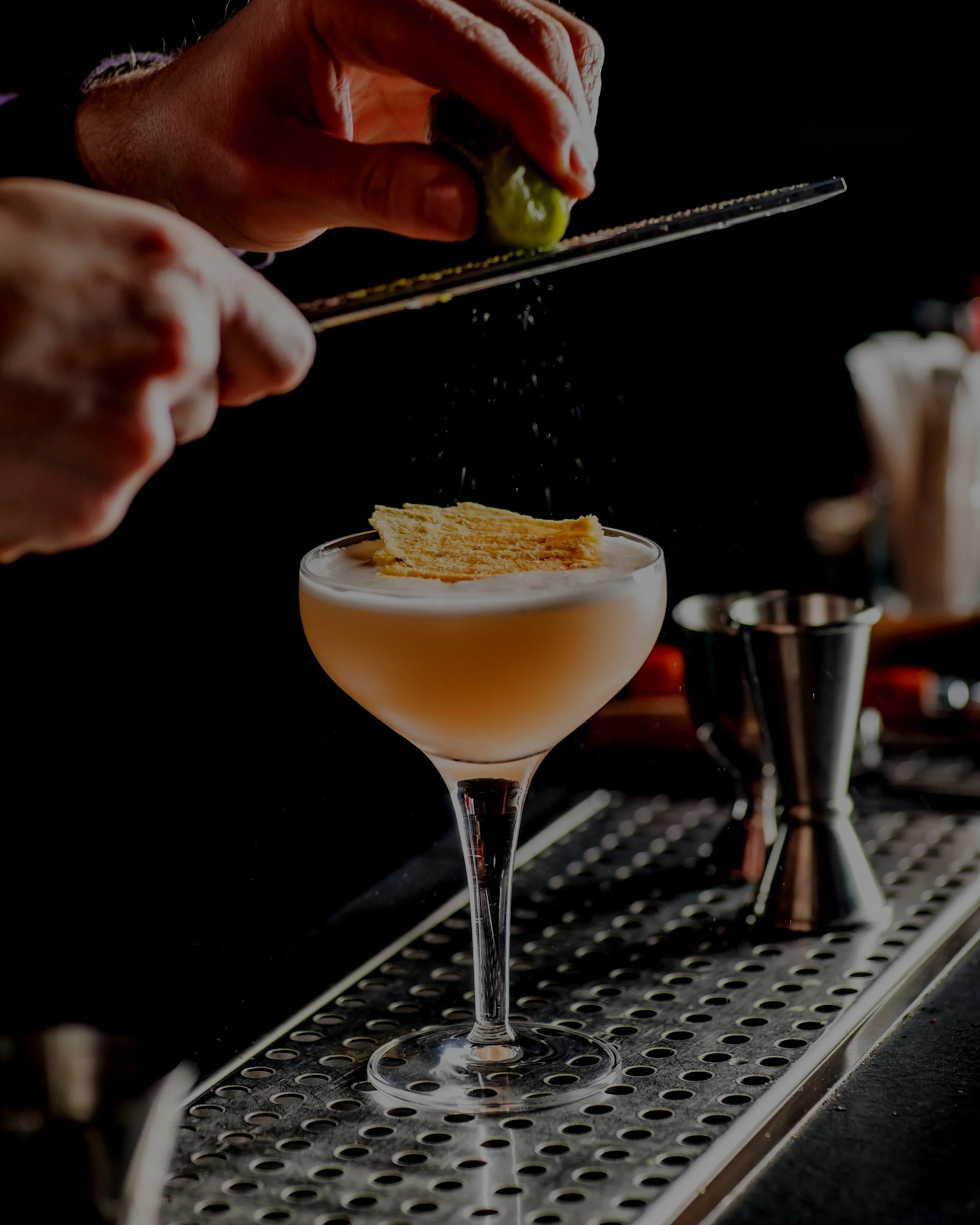 A bartender's hand is squeezing a lime wedge over a cocktail in a coupe glass, which has a salted rim and a garnish of a lime wedge on top.
