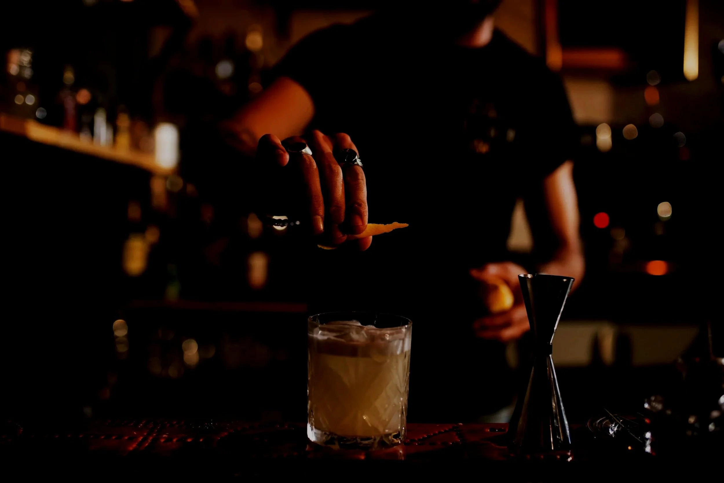 A person preparing a cocktail drink in a dimly lit bar with a blurred cityscape background.