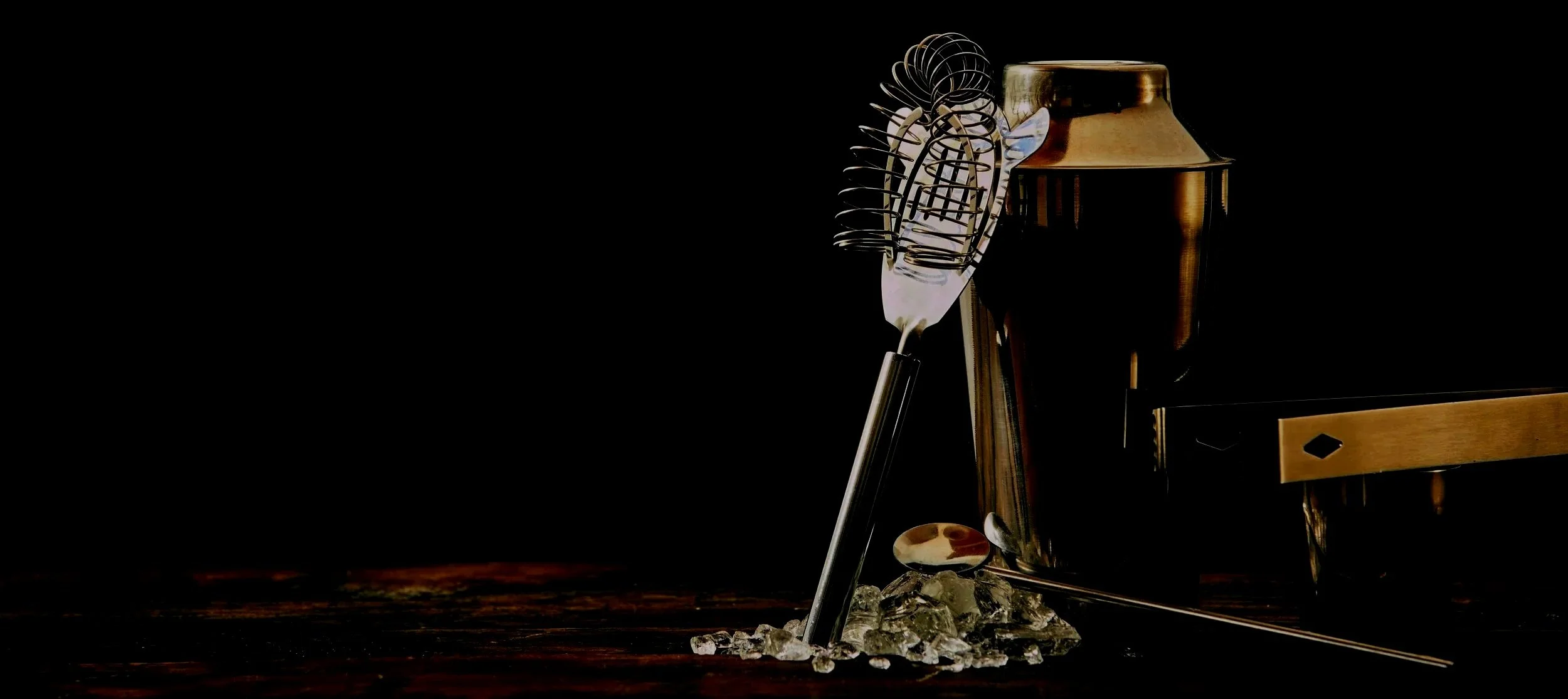 Bar tools including a cocktail strainer, shaker, and ice on a dark wooden surface.