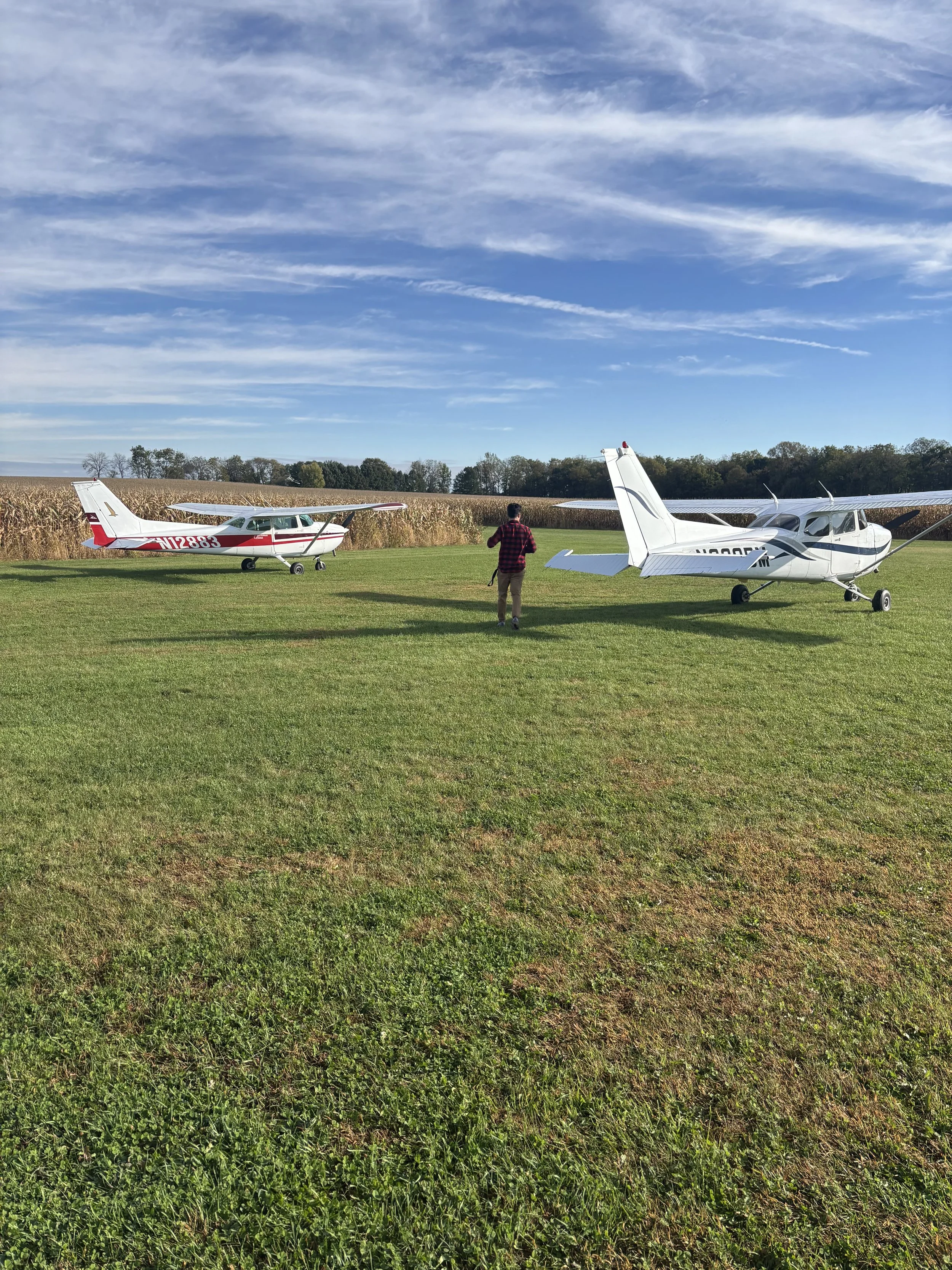 A person in a red and black plaid shirt walking between two small airplanes on a grassy field with farmland and blue sky in the background.