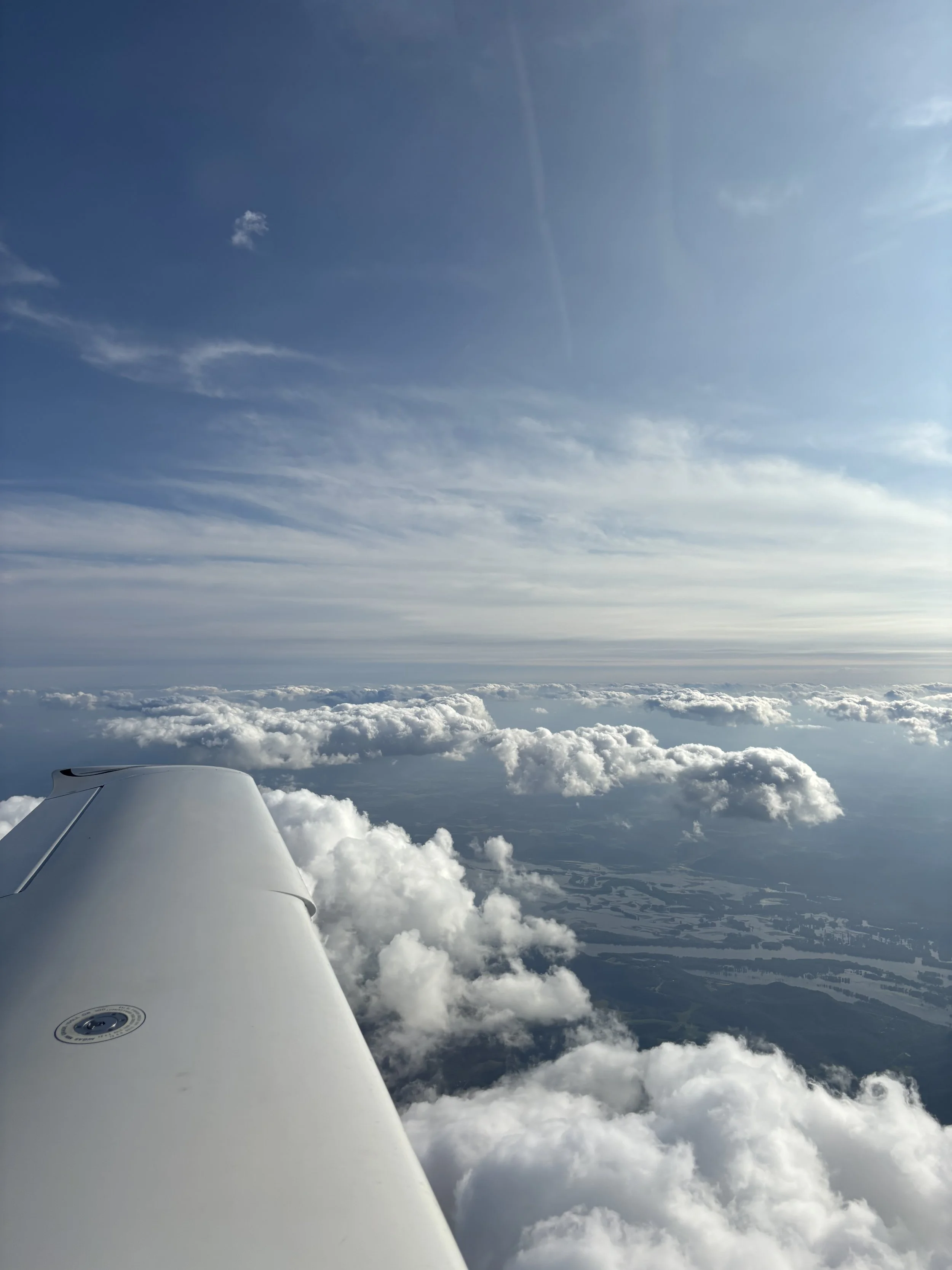 Photo taken from an airplane window showing part of the airplane wing, with fluffy clouds and a bright blue sky above and below. Some land and water bodies are visible below.