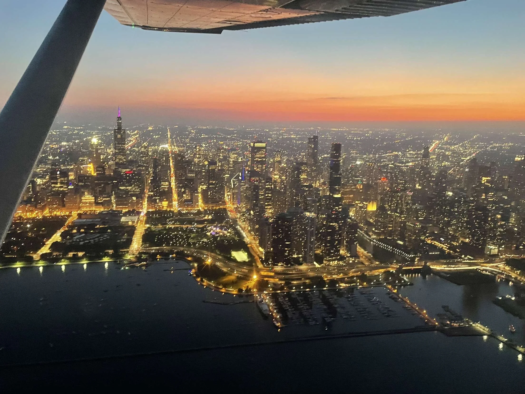 An aerial view of New York City skyline at sunset, seen from a helicopter, with tall skyscrapers, city lights, and a river visible.