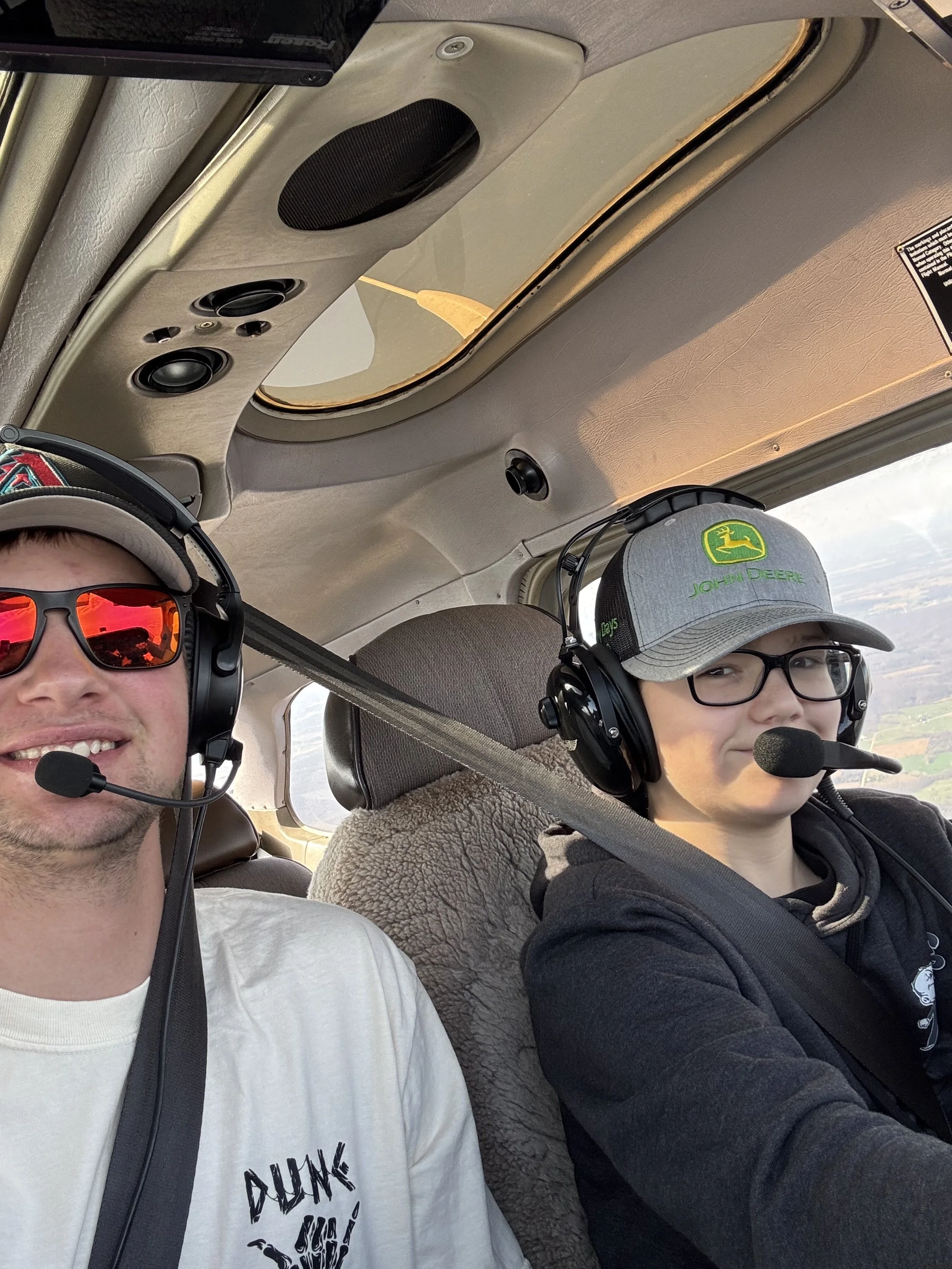 Two people wearing headsets and sunglasses sitting in the cockpit of a small aircraft, with a view of the sky and landscape through the window.