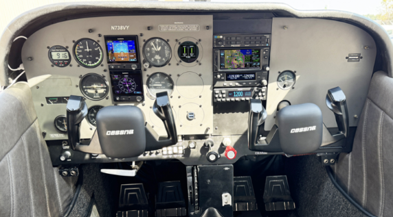 The cockpit of a small aircraft featuring various gauges and avionics, with two control yokes marked 'CESSNA', and a central instrument panel.